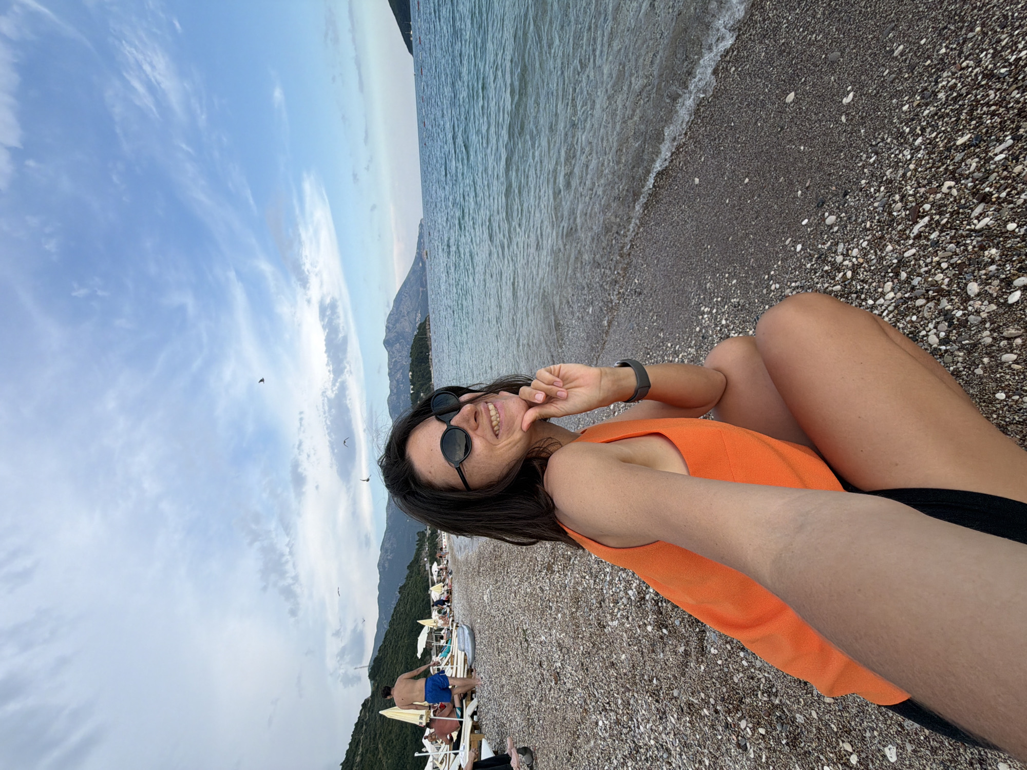 A woman wearing an orange tank top and black shorts squats on a pebble beach, holding her chin with her right hand. She wears round, black sunglasses and smiles at the camera. The sea is to her right, with gentle waves lapping the shore. In the background, a beach with lounge chairs, umbrellas, and several people is visible, backed by a hillside covered in trees and mountains under a cloudy sky with several birds flying. This scene is on a beach in Budva, Montenegro.