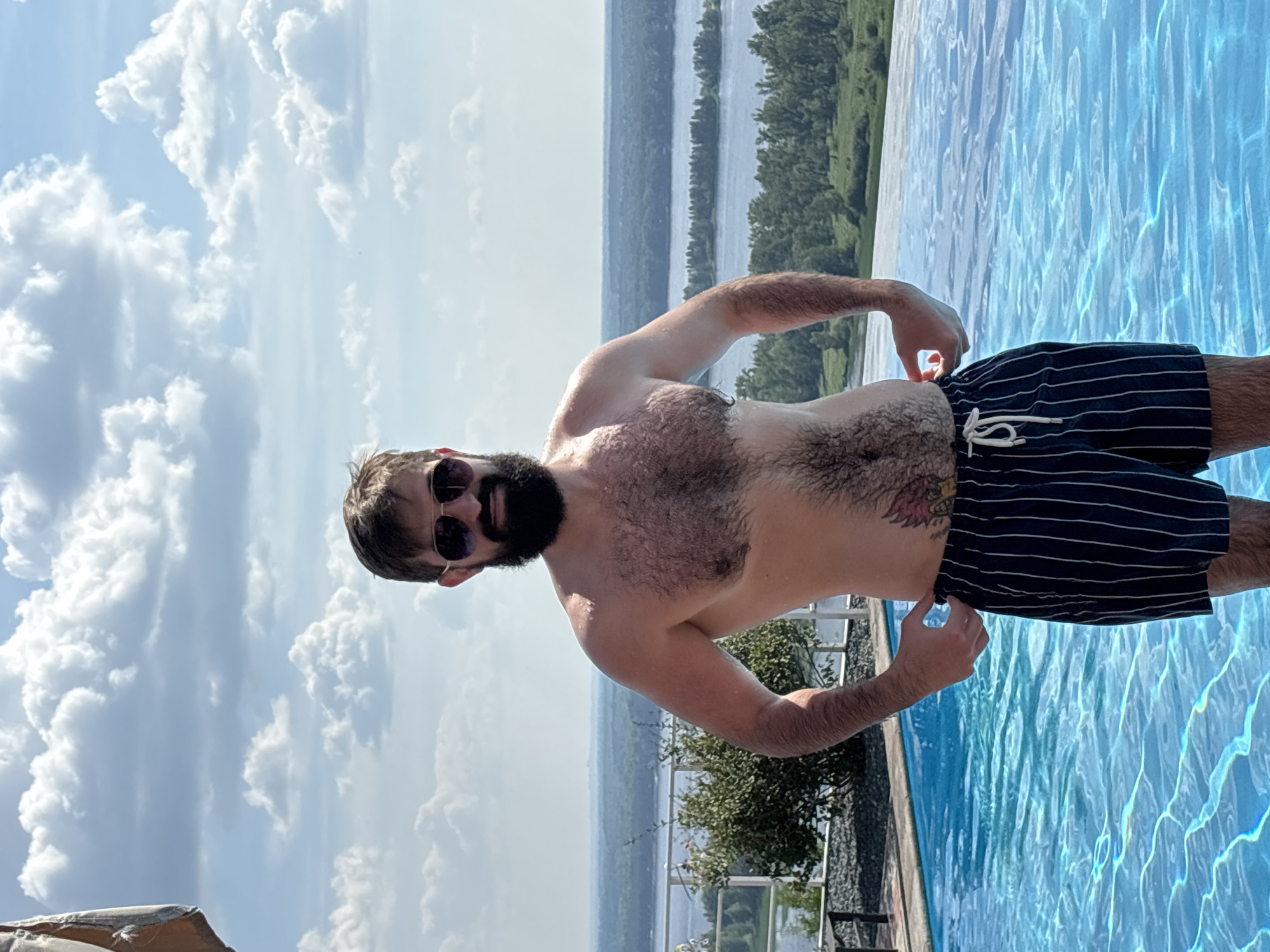A man with a beard and sunglasses stands near an infinity pool. He is wearing dark swim trunks with white pinstripes. The pool has clear blue water that reflects the sunlight. Behind the pool, there is a body of water and lush greenery. The sky is bright blue with fluffy white clouds, indicating a sunny day. The man is looking towards the camera with a relaxed expression. A tattoo is visible on his side. The setting appears to be a resort or a private residence with a scenic view.