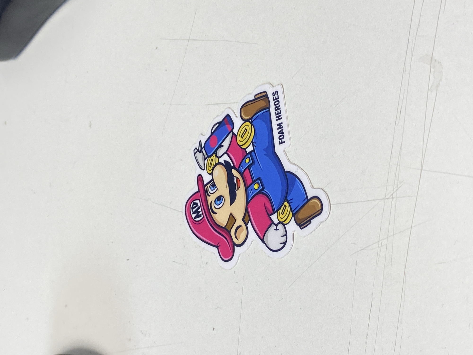A sticker depicting a cartoon character resembling Mario, wearing a pink hat with "MD" on it, a pink shirt, and blue overalls, is placed on a white surface. The character is holding a blue spray bottle with pink accents. The sticker has the text "FOAM HEROES" printed on it. The white surface has faint lines and specks on it. No information about the city of Uchaly, Russia, is present in the image.