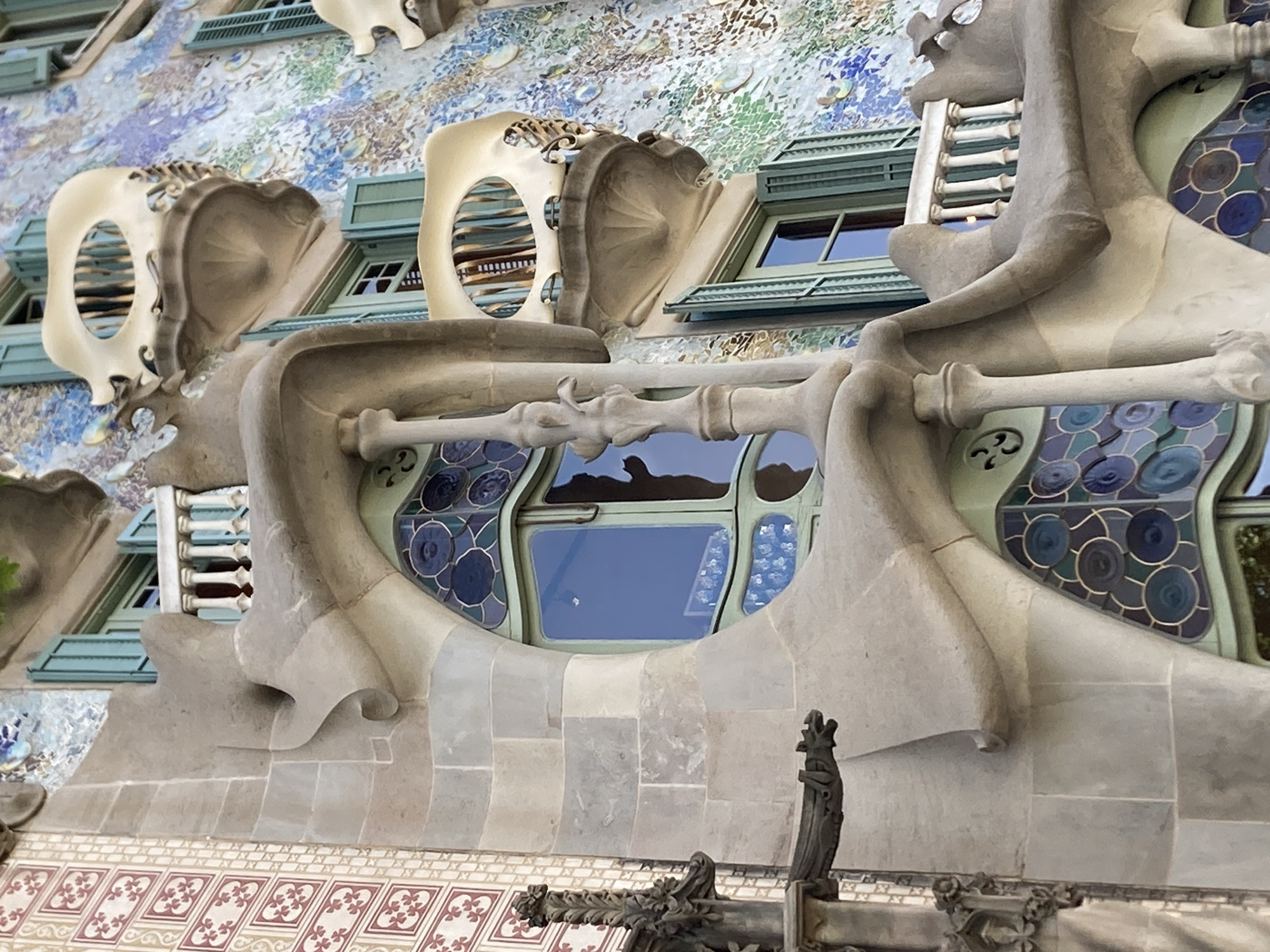 The image shows a close-up, exterior view of a section of Casa Batlló, a famous building located on Passeig de Gràcia in Barcelona, Spain. The architecture is characterized by its organic, flowing lines and vibrant mosaic facade.
On the left, a section of the building's facade is constructed from large, pale grey stone blocks with visible mortar lines. This stone section features a prominent, curved protrusion with a scrolled end, resembling a bone-like structure. To the far left, a decorative, gothic-inspired gargoyle figure is attached to the stone facade. Adjacent to the stone, on the upper left, is a vertical band of decorative tiling with a repeating pattern of red floral motifs on a cream background.
The central and right portions of the image showcase the building's colorful mosaic work, composed of numerous small, irregularly shaped tiles in shades of blue, green, white, and yellow, forming a shimmering, almost iridescent surface. Interspersed within this mosaic are several distinctive balconies. These balconies are made of a creamy, bone-white material and have an unusual, biomorphic shape, resembling masks or stylized faces. Each balcony has an oval opening with thin, vertical bars behind it, suggesting a balustrade.
Several windows are visible. Some have light blue or teal shutters, which are partially open. One large window in the center is framed by the stone architecture and features a dark blue reflective glass. Below this window, and visible in the lower half of the image, are sections of stained glass with a circular, kaleidoscopic pattern in shades of dark blue and teal, framed by green and gold lines. A white balustrade with vertical spindles is visible along a balcony on the right side of the image. The overall impression is one of whimsical, surreal, and highly decorative architectural design.