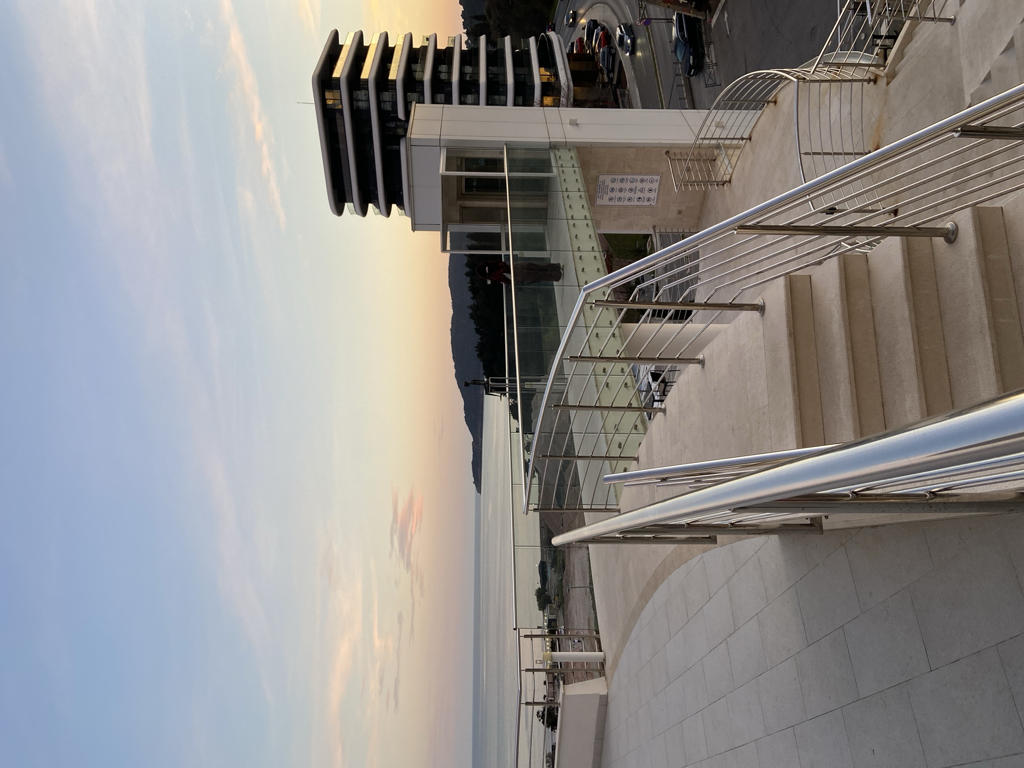 An elevated viewpoint in Boreti, Montenegro, captures a coastal scene at dusk, characterized by the warm, fading light of a sunset. The sky displays hues of light blue, orange, and soft pink with wispy clouds.
In the foreground, a light-colored tiled patio curves to the left, while a set of stone steps with modern stainless steel handrails featuring horizontal bars ascends into the frame from the lower right. These stairs lead to an elevated observation deck or walkway.
The observation deck, constructed with glass and stainless steel railings, extends towards the sea. One individual, appearing to be an adult in dark clothing, stands on this deck, facing away from the viewer, observing the distant horizon. An enclosed glass elevator shaft is integrated into the structure adjacent to the deck. Below the observation deck, a sign displaying multiple pictograms (e.g., 'no smoking', 'no pets') is affixed to the building's facade.
To the right, a multi-story, modern building with curved, dark-colored balconies and reflective windows rises prominently, illuminated by the low sun. A curved asphalt road runs at the base of this building, occupied by several parked and moving vehicles, including a dark sedan and what appears to be an SUV.
The background features the calm, expansive surface of the Adriatic Sea, stretching to the horizon. A distant coastline with hills covered in green vegetation is visible. Closer to the shore, several low-rise structures and a beach area are identifiable.