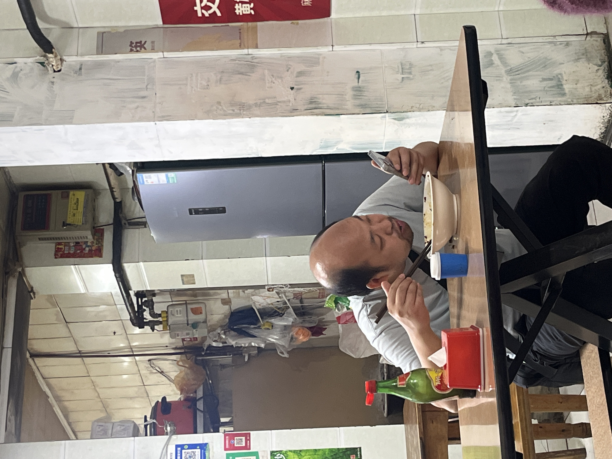 In Jiulongpo District, China, a person sits at a wooden table in a brightly lit, somewhat cluttered interior. The individual, balding with dark hair, is using chopsticks to eat from a white bowl while simultaneously holding a smartphone in their left hand. A green bottle with a red cap and a red tissue box sit on the table. A blue cup is also visible. Behind the person, a stainless steel refrigerator can be seen against a tiled wall. Overhead, a complex network of pipes and fixtures is visible. Several posters or advertisements are on the wall.
