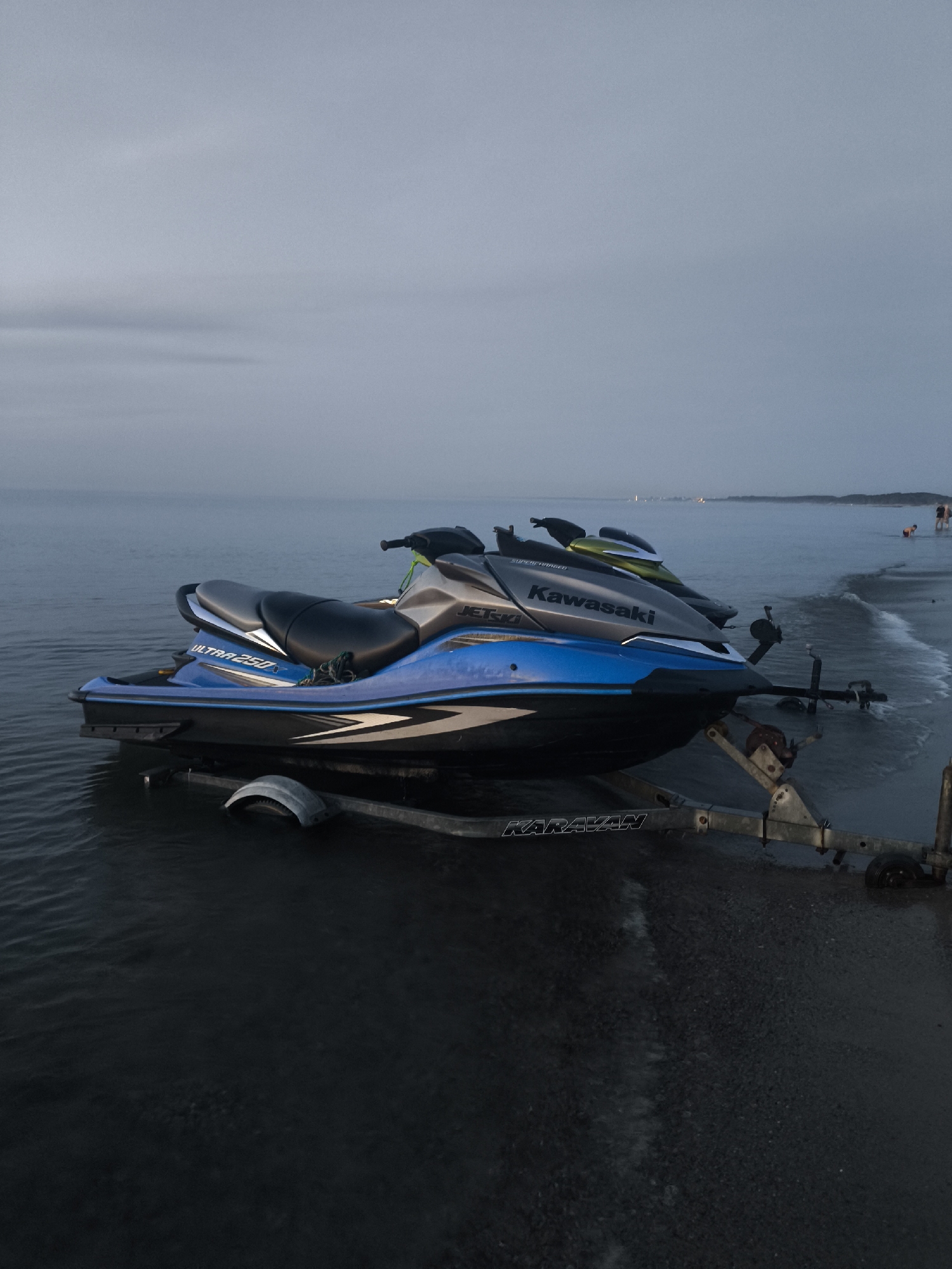 The image depicts a scene at the beach in Куликово, Russia, near the water's edge. Two jet skis are positioned on a trailer at the beach, with the front jet ski partially submerged in the shallow water. The jet skis are a bright blue and silver, with "ULTRA 250" and "Kawasaki" visible on their sides. The trailer is silver and has visible wheels. In the background, a hazy horizon separates the water from the sky, which is overcast and a light gray color. There are a few people visible in the distance on the beach and in the water, appearing as small, indistinct figures. The overall scene is serene and appears to be during twilight or an overcast day.