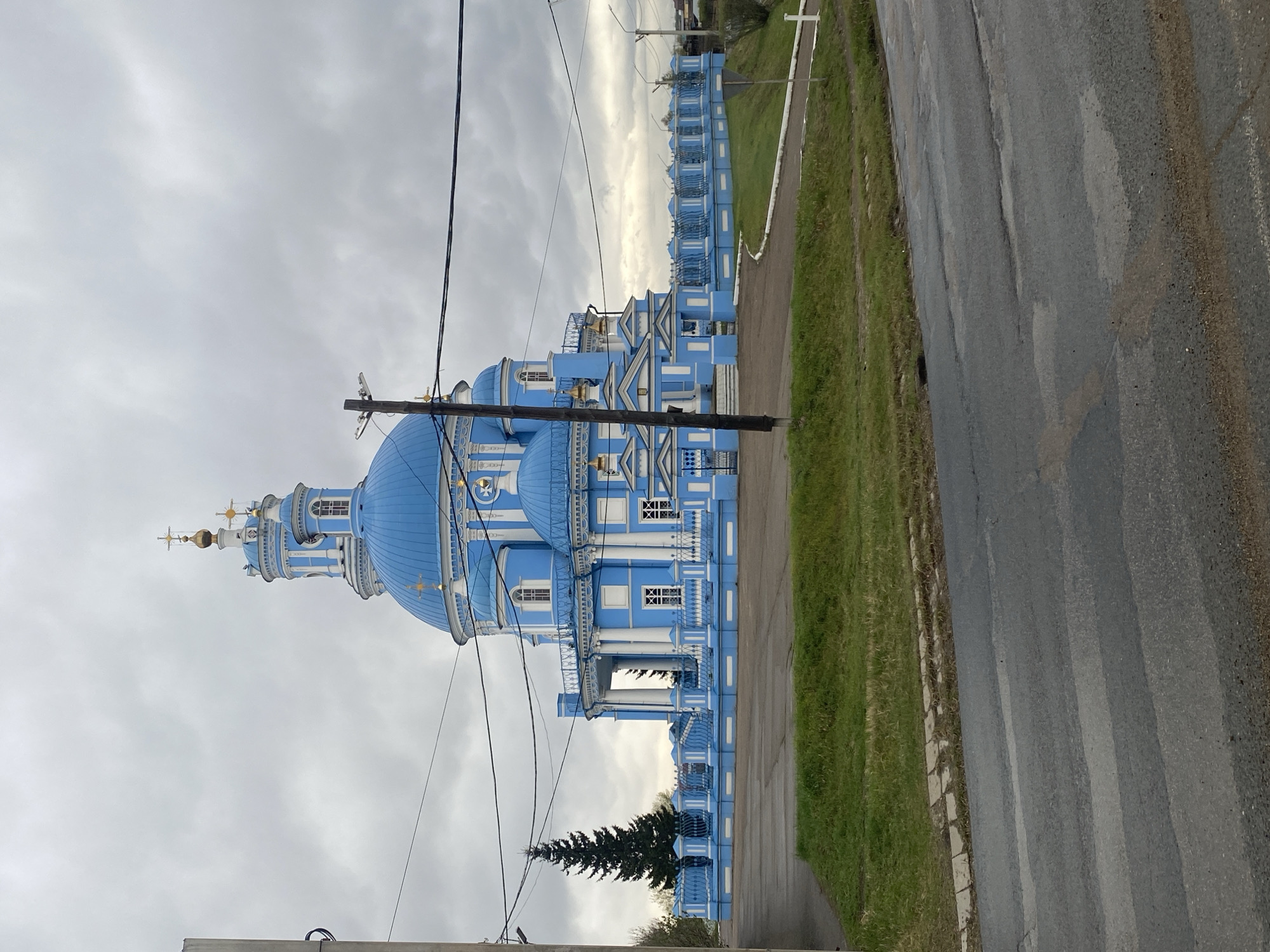 The image depicts the Church of the Kazan Icon of the Mother of God in Telma, Russia. The church is primarily blue and white with multiple domes. It is situated near a road and a grassy area. Power lines are visible in the foreground. The sky is overcast.