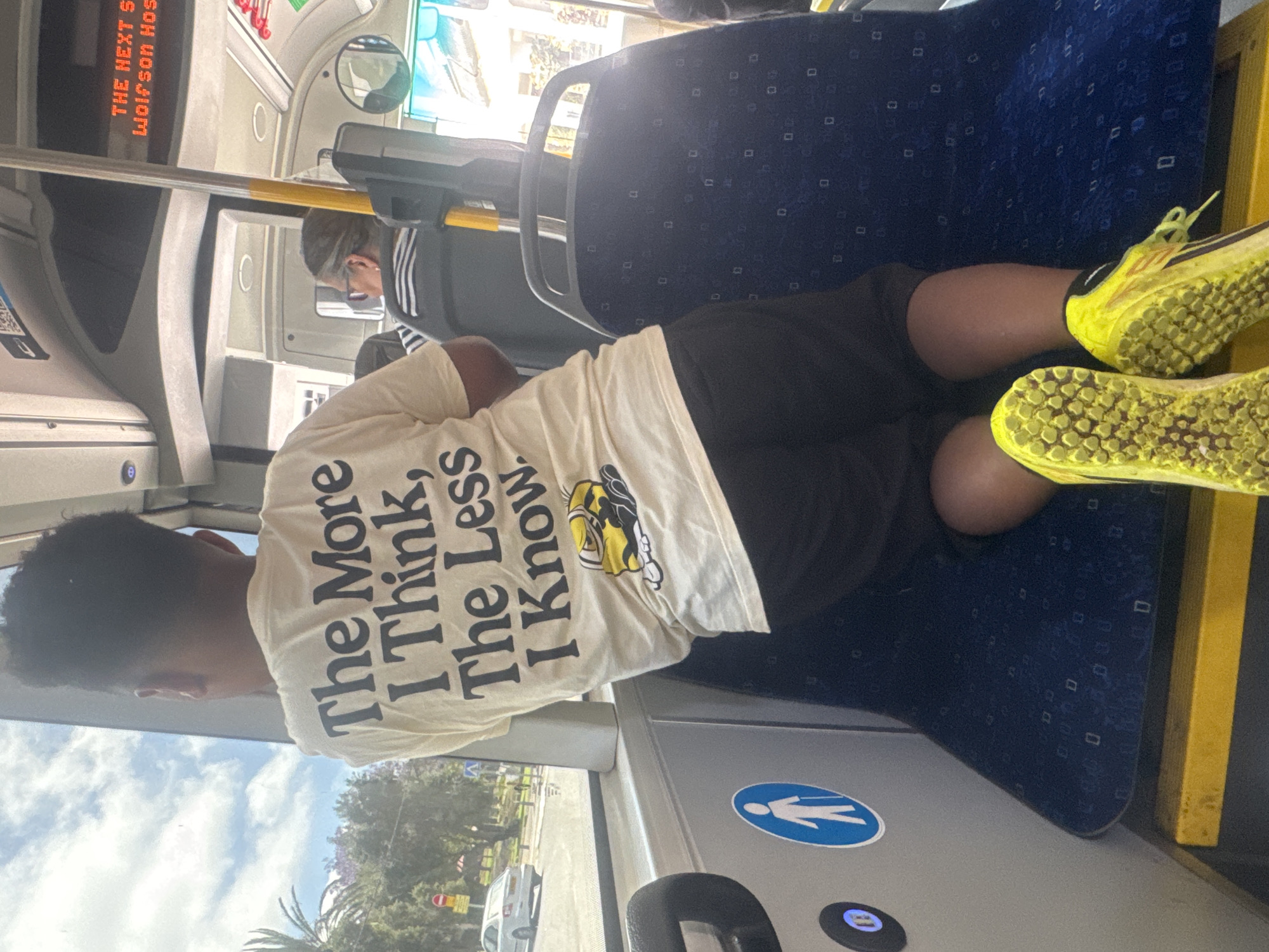 The image depicts a person lying horizontally across seats on a public bus in Tel Aviv, Israel. The person is wearing a white t-shirt with the phrase "The More I Think, The Less I Know," black shorts, and bright yellow athletic shoes. The person is positioned against the blue patterned seat. Another person is partially visible further up the bus. The interior of the bus is visible, with handrails and destination display showing the "Wolfson Hospital". Outside the bus window, there is a street with trees and vehicles.