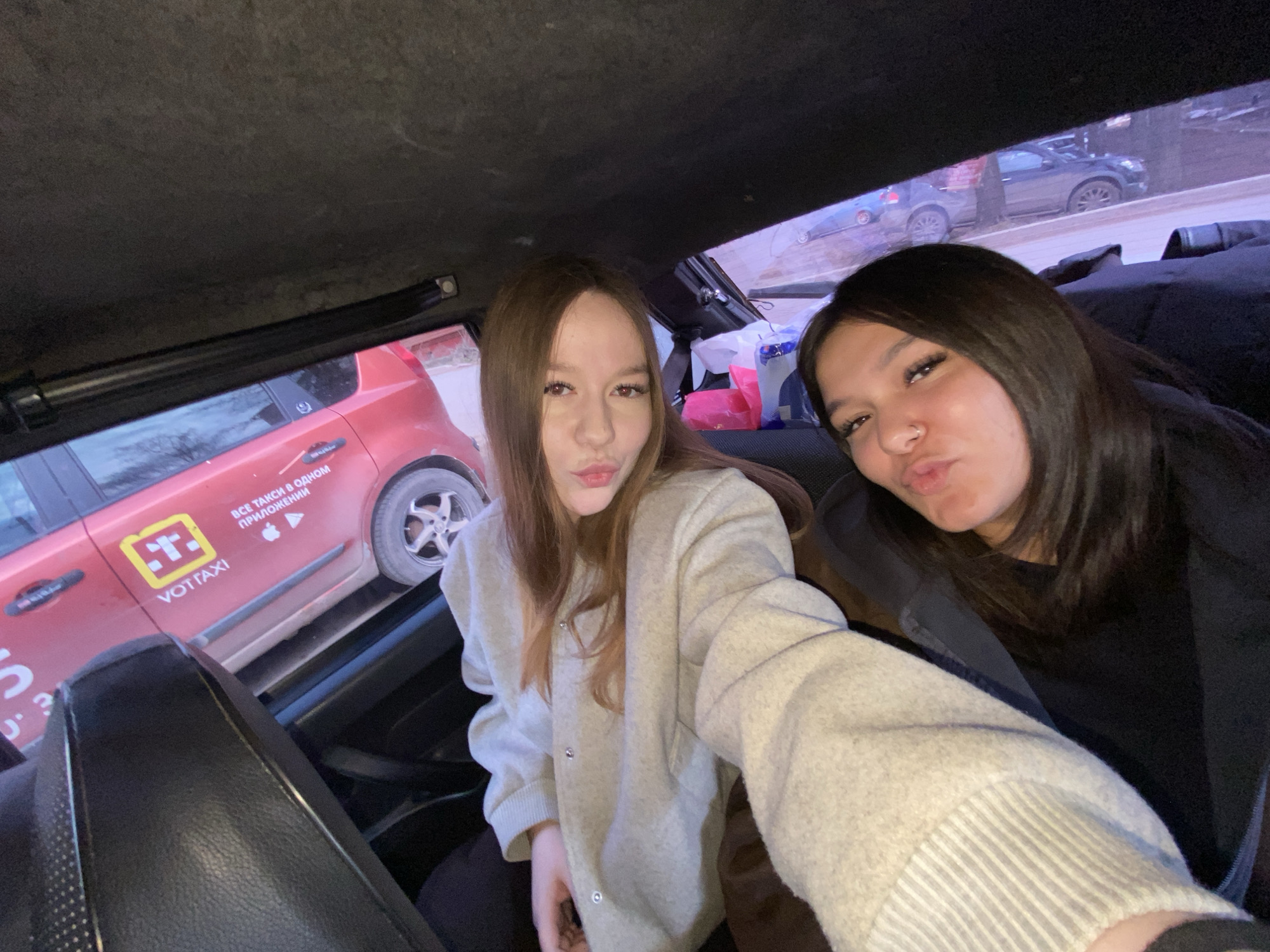 Two women are sitting in a VOTTaxi car in Votkinsk, Russia. The car is red and the taxi company’s logo is visible on the side. The women are making duck-lips toward the camera. One woman is wearing a light gray jacket and the other is wearing a black shirt. Outside of the car, other cars and trees are visible.
