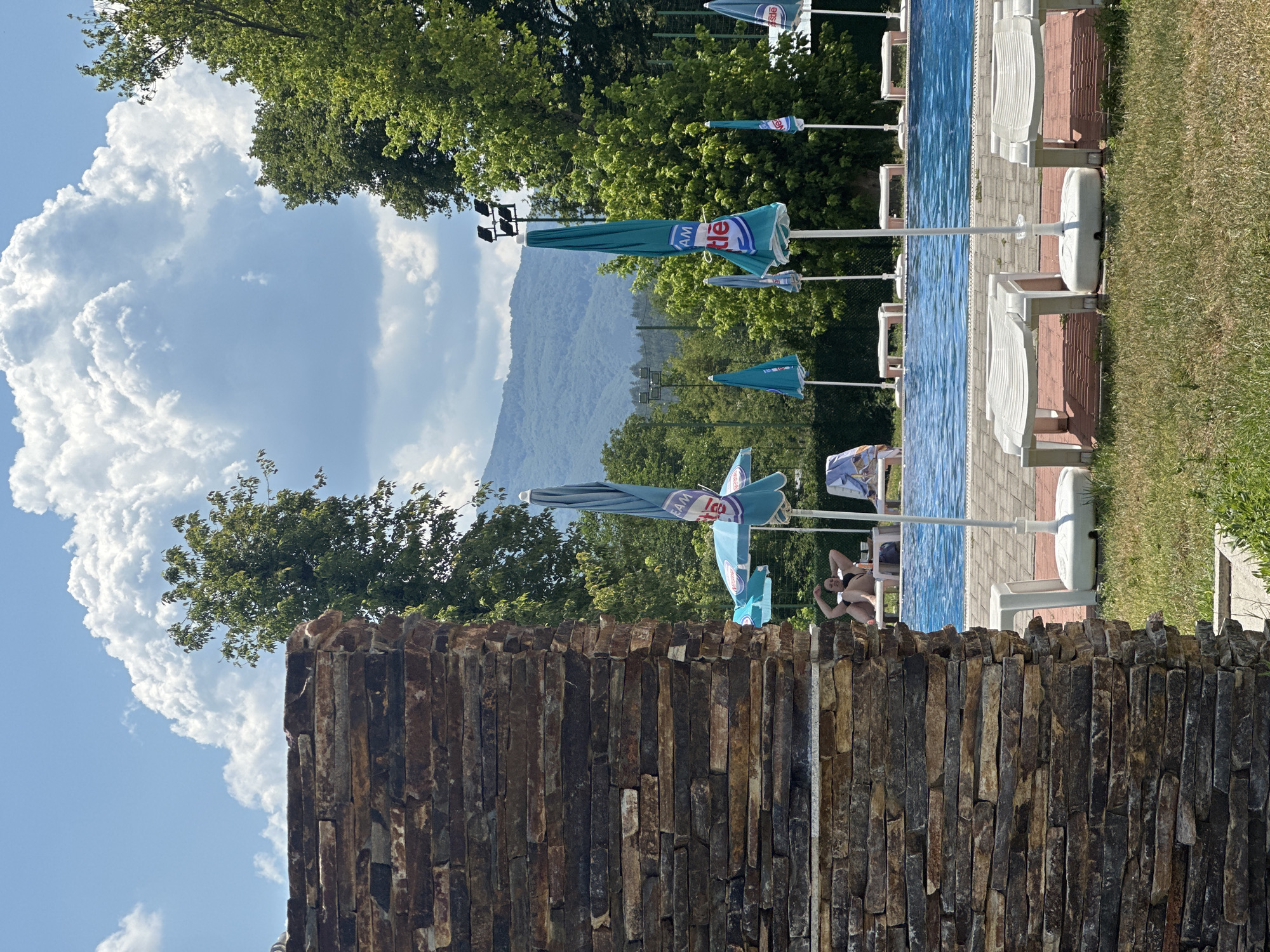 In Simitli, Bulgaria, on a sunny day with a large cumulonimbus cloud formation in the sky, a scene unfolds by a swimming pool. A stone wall, constructed from irregularly shaped, multi-colored stones, dominates the left foreground. Beyond this wall, a vibrant blue swimming pool is visible, with the water reflecting the bright sky.
Along the poolside, several white lounge chairs with patterned cushions are arranged. Scattered around the pool area are closed turquoise umbrellas, some bearing the "Nestle" logo, indicating a sponsorship or branding. A few people are present in the scene; in the middle ground, a person is relaxing on a lounge chair near the pool, their arms raised. In the background, lush green trees frame the pool, and further back, rolling hills and mountains are visible under the vast sky. The ground around the pool is a mix of green grass and a paved area with patterned bricks. The lighting suggests it is daytime, likely late morning or early afternoon.