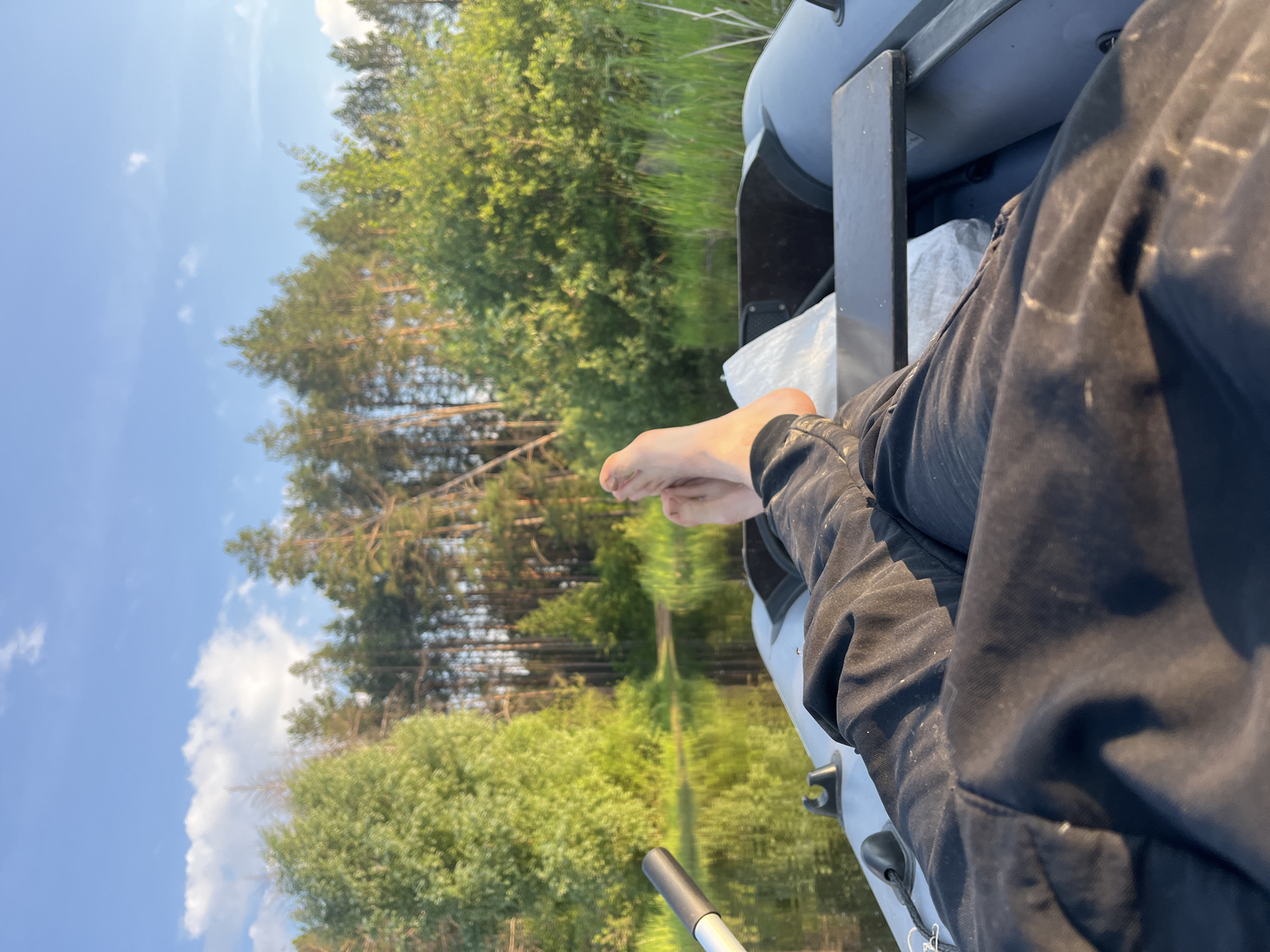 A first-person perspective shot shows a person's bare feet and legs in black pants, resting in an inflatable boat on a calm body of water. The boat's gray interior is visible, with an oar handle to the left and a white bag further inside. The water reflects the surrounding lush green trees and a clear blue sky with scattered white clouds. The background consists of a dense forest of tall pine trees and other green foliage lining the water's edge. The scene suggests a peaceful outdoor activity, likely a boat ride on a lake or river, possibly in or near Chaikovsky, Russia, given the typical landscape.