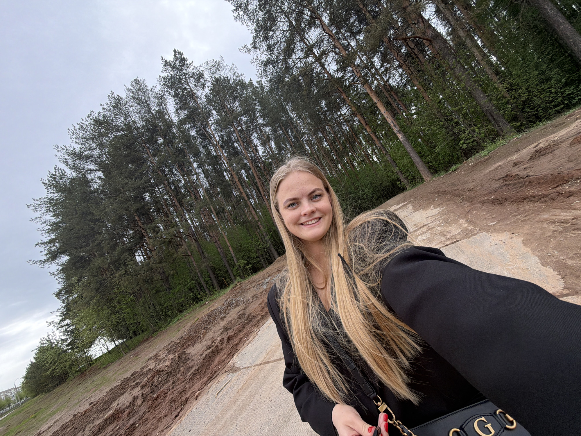A young, fair-skinned blonde woman is taking a selfie outdoors. She is smiling at the camera. She is wearing a black blazer and holding a black purse with gold details. The location appears to be a park or wooded area on the outskirts of a city. In the background, there's a dirt patch on the left, and a dense forest of tall trees with green foliage extends upward towards an overcast sky. The image was taken in Izhevsk, Russia.