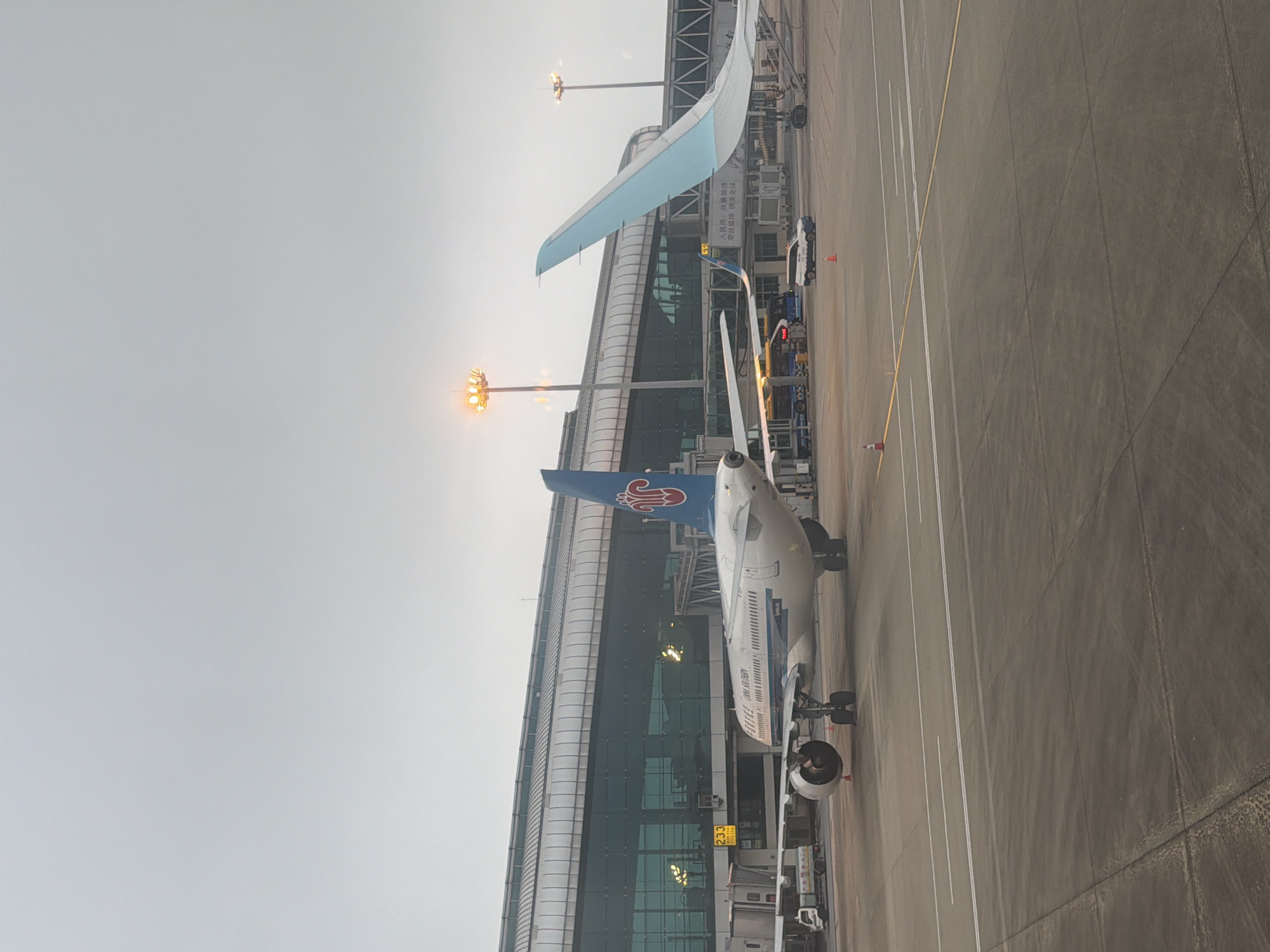 The image shows a China Southern Airlines aircraft parked at a gate at Guangzhou Baiyun International Airport in Baiyun District, Guangzhou, China. The aircraft is positioned adjacent to a terminal building with glass windows. Ground crew vehicles and equipment are visible near the plane. The sky is overcast.