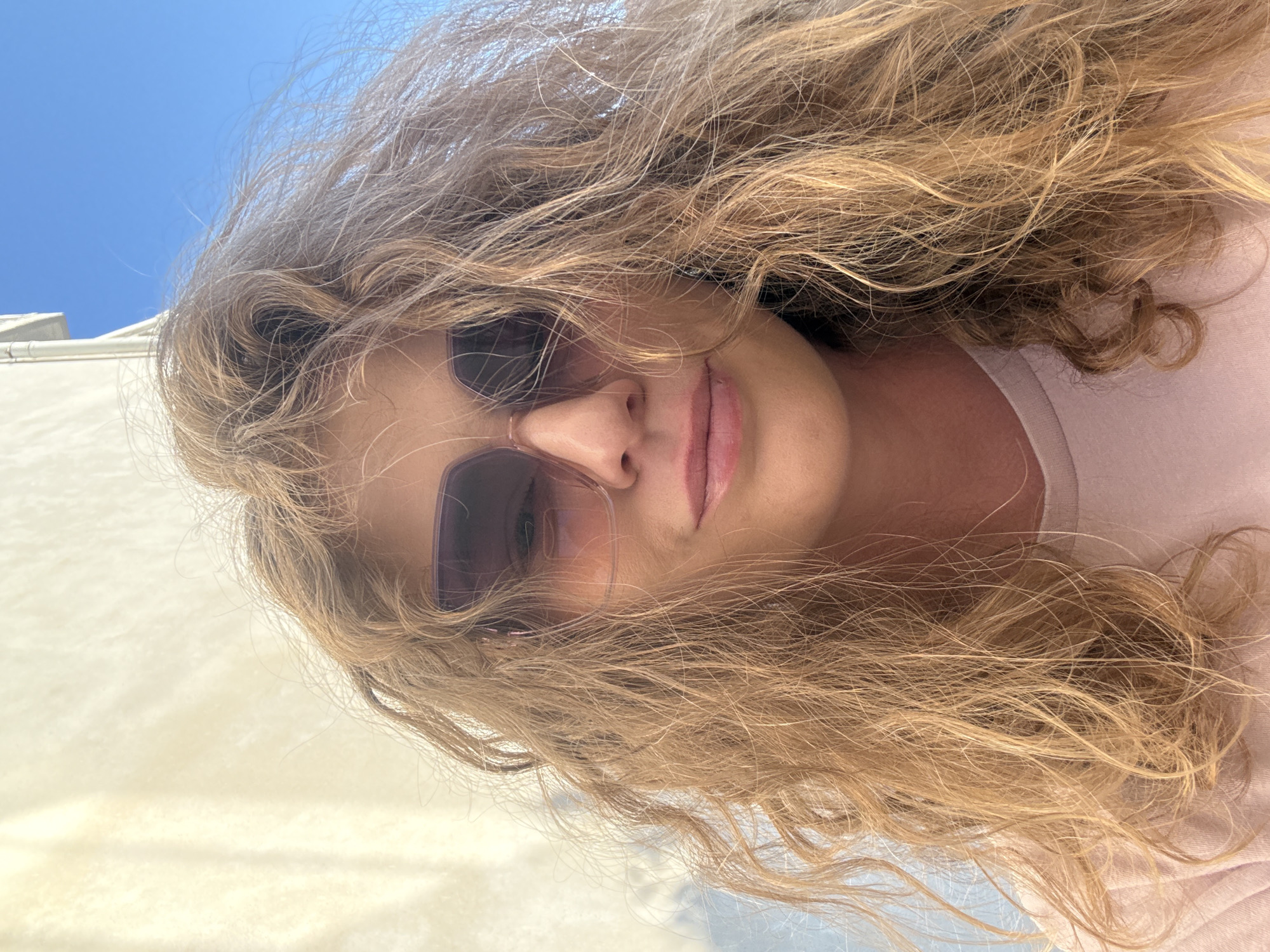 A woman with curly, light-brown hair is wearing sunglasses and a light-colored shirt. The background shows a glimpse of blue sky and white structures, possibly part of a building or an outdoor setting. The location is inferred to be in Budva, Montenegro.