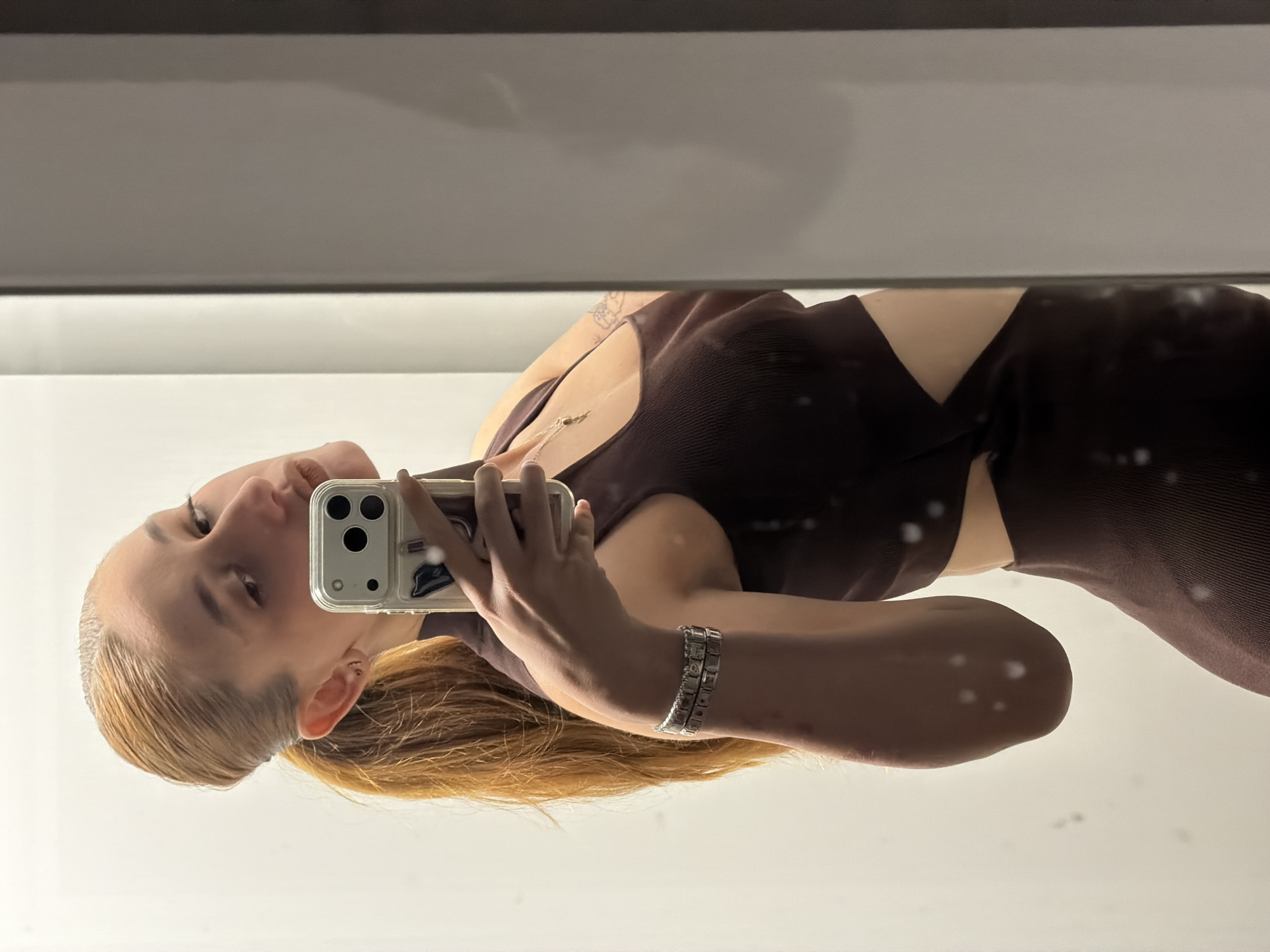 A young woman with blond hair tied back, wearing a brown dress with side cutouts, is taking a selfie in a mirror. She is holding a silver-colored smartphone in her left hand, with a silver bracelet visible on her wrist. The setting appears to be indoors, likely a dressing room or bathroom, with neutral-colored walls and lighting. The location in Plovdiv, Bulgaria, is not discernible from the image.