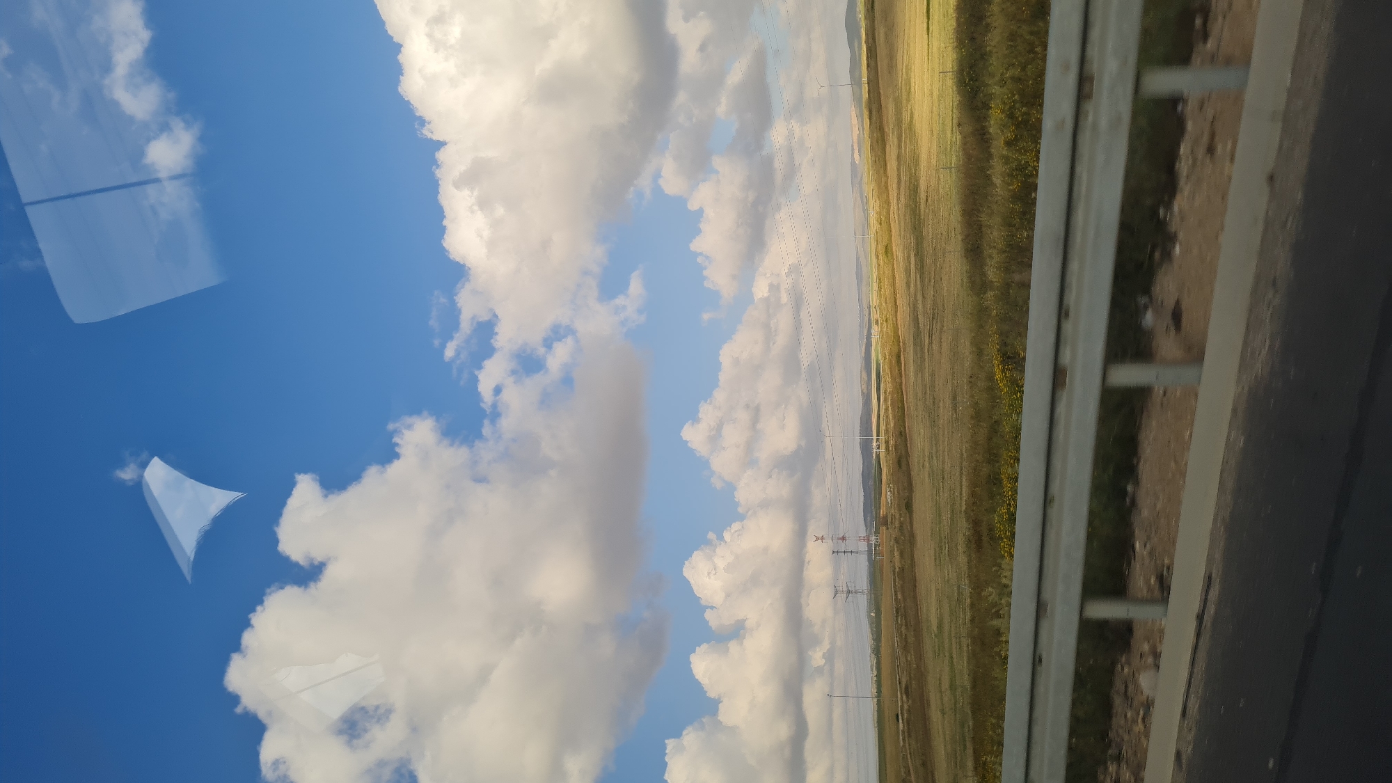 The image, taken from inside a vehicle in the Bnei Shimon Regional Council, Israel, shows a landscape of a field under a partly cloudy blue sky. Reflections are visible on the glass, obscuring portions of the view. A metal highway guardrail is prominent in the lower right. Power lines and towers are visible in the background. The field appears to be dry grassland, with some yellow wildflowers. A road is visible in the foreground along with the guardrail.