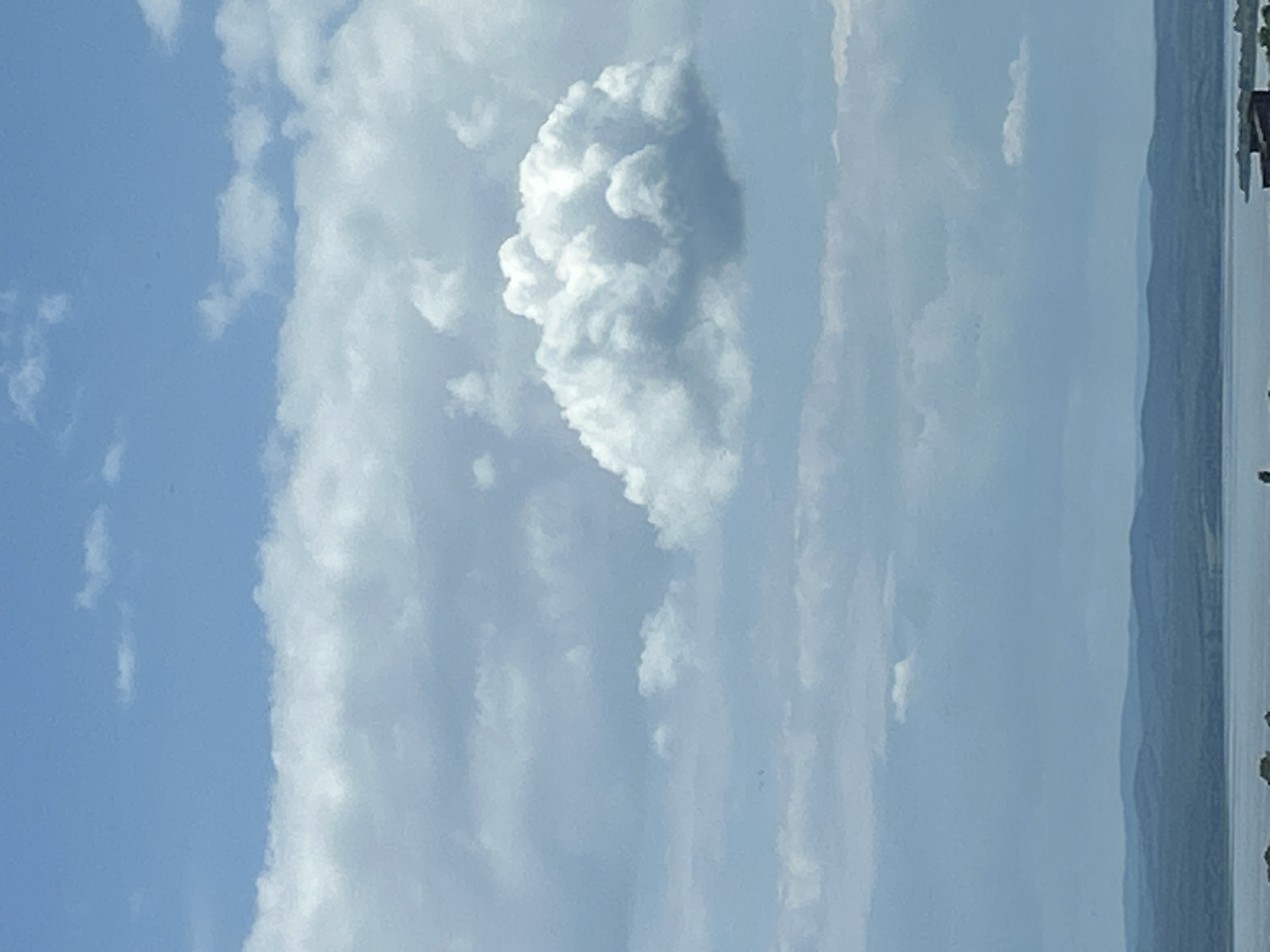 The image is a vertical shot of a cloudy sky, possibly in Burgas, Bulgaria. Fluffy white clouds of varying sizes occupy the center and left of the frame, set against a blue sky. On the right side, there are land formations, seemingly distant hills or mountains. The edge of a body of water, perhaps a lake or the sea, is also visible on the extreme right, with some indistinct structures or vegetation along the shore. The image appears to have been taken during the daytime in outdoor setting.