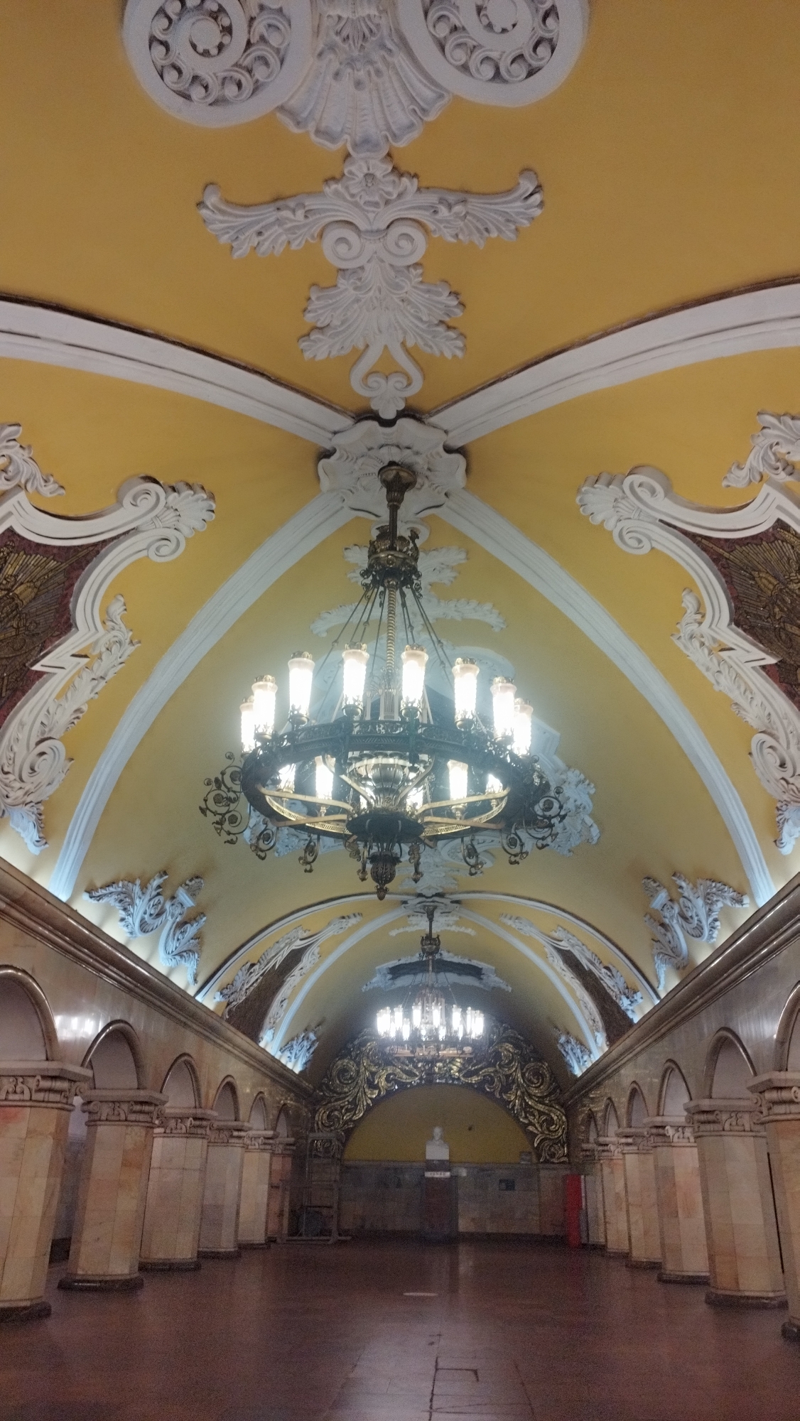 The image shows the interior of a grand metro station in Moscow, Russia, likely part of the Moscow Metro system, known for its ornate architecture. The ceiling is a prominent feature, painted a bright yellow and adorned with elaborate white stucco moldings and intricate decorative elements. A large, multi-tiered chandelier with numerous lights hangs from the center of the arched ceiling, casting a warm glow.
The station features a long, wide hall with arched openings supported by fluted marble columns on either side. The walls are paneled with marble or a similar stone, giving a sense of opulence. In the distance, at the far end of the hall, another chandelier is visible, illuminating an alcove that features a bust on a pedestal, set against a backdrop of ornate golden stucco work. The floor is a polished, dark material that reflects the light. The overall impression is one of grandeur and historical significance, characteristic of many Moscow Metro stations. There are no people visible in the image.