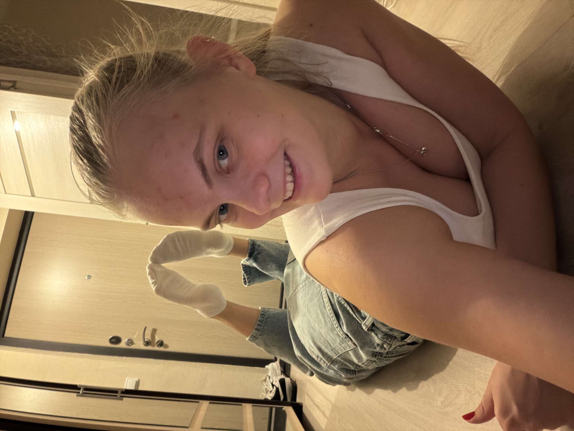 A young woman is lying on a light-colored floor. She is wearing a white tank top and denim shorts. Her feet, clad in white socks, are raised in the air behind her. She is smiling at the camera. In the background is a light-colored door. A pair of shoes can be seen near the bottom of the frame.
The image does not contain any information regarding the city of Izhevsk, Russia.