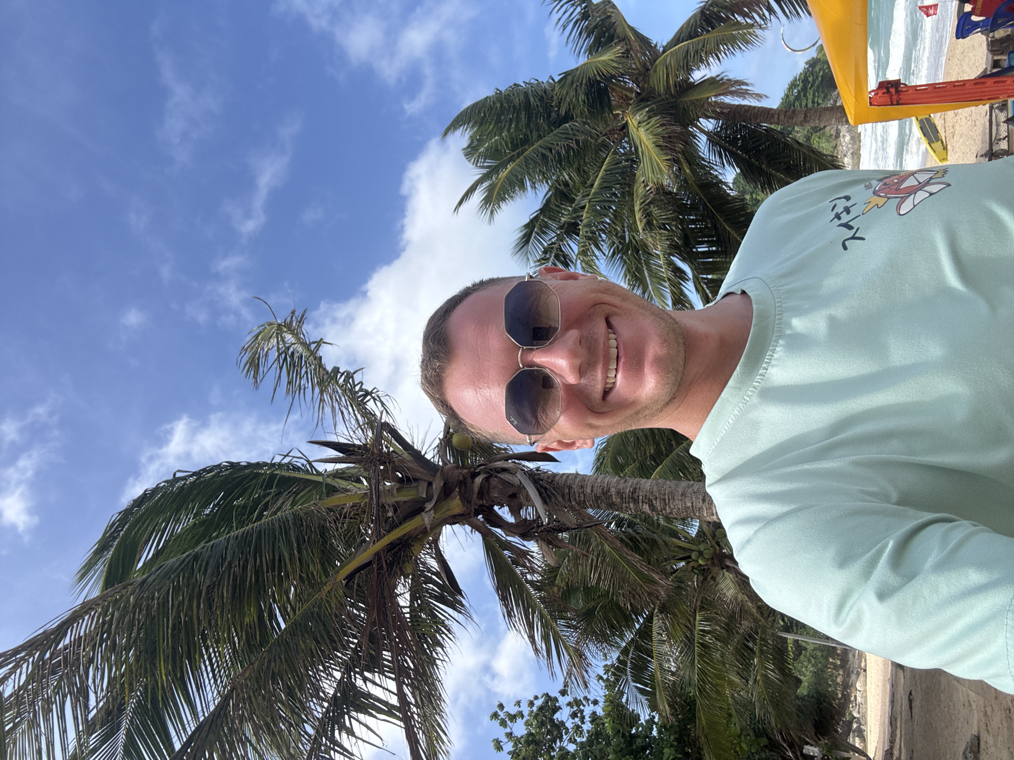 A man takes a selfie on a beach in Rawai, Thailand. He is wearing sunglasses and a light green t-shirt with a Japanese character and a cartoon image of a fish. Behind him, palm trees sway under a blue sky with scattered clouds. The beach is sandy with some vegetation and a yellow structure visible to the right. The ocean and waves are also visible in the background.
