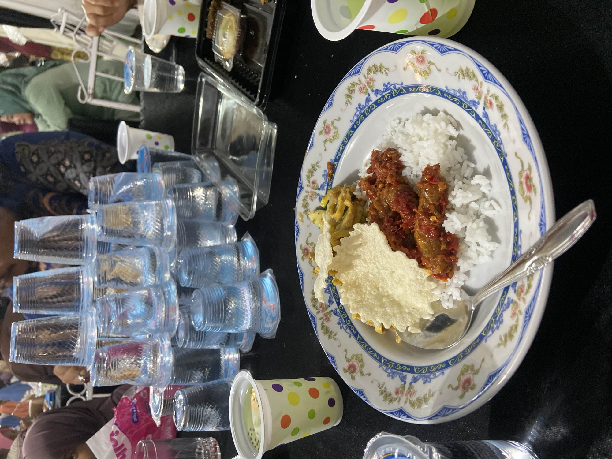 The image depicts a table laden with food and drinks, likely during a meal or gathering in Air Molek II, Indonesia. The scene includes stacks of clear plastic cups, some filled with water and others empty. One prominent object is a white plate with a decorative blue and floral rim, filled with white rice, a reddish-brown dish likely containing meat in sauce, and a piece of crispy cracker or wafer. A silver spoon rests on the plate. To the left, there are several clear plastic containers, one of which holds what appears to be a dessert or pastry. In the background, partially visible, are people. One person is wearing a light green headscarf and a dark top, and another is wearing a dark blue headscarf with a patterned fabric. The setting appears to be indoors, with a dark tablecloth covering the table. There are also some colorful paper cups with polka dot patterns. The lighting suggests it could be daytime or artificial lighting, but no specific time of day or weather can be determined. No discernible text is visible in the image, apart from what might be branding on a pink plastic bag in the lower left corner, which is too blurry to read. The overall impression is one of a communal meal, possibly a celebration or a regular gathering.