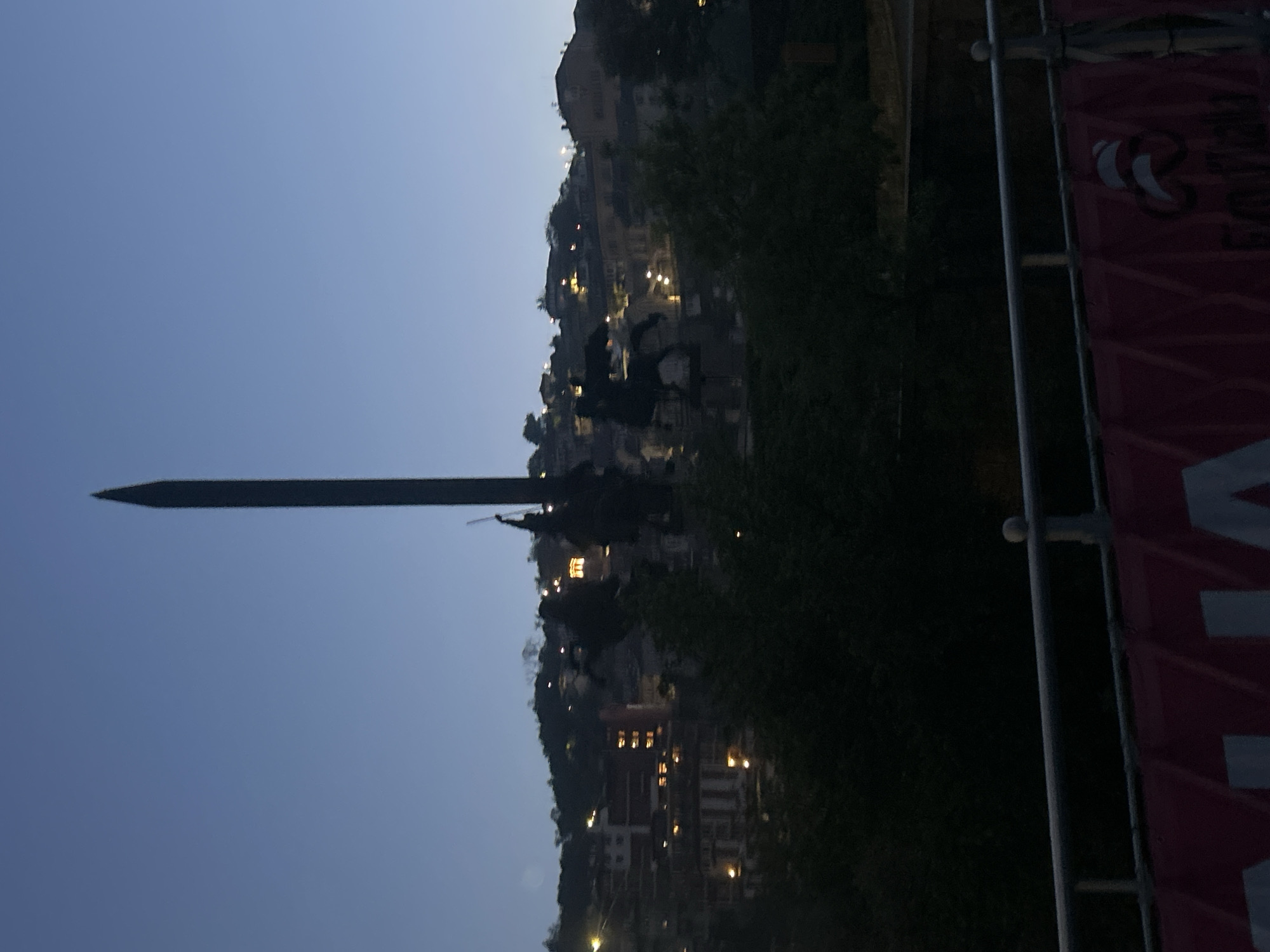 The image shows a view in Veliko Tarnovo, Bulgaria, at dusk. The Baldwin monument with a long, tapering spike atop a male equestrian figure is visible against the evening sky. Below the monument is the cityscape illuminated by scattered lights on a hillside. The right side of the frame includes the edge of a dark banner with a partial logo visible against a supporting metal frame. Trees fill in the background around the hillside.