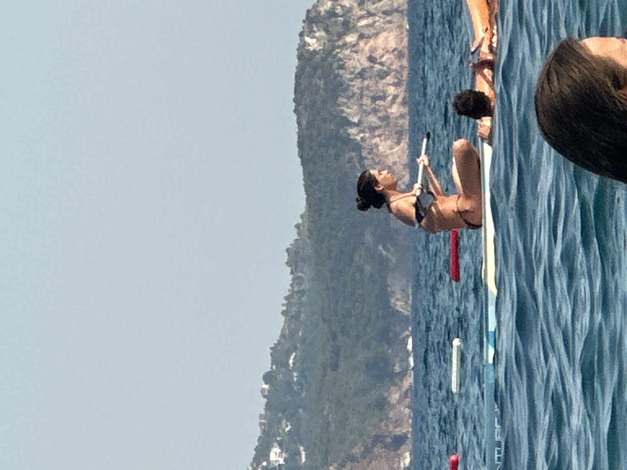 In Boreti, Montenegro, a woman in a bikini sits on a paddleboard in the Adriatic Sea, holding a paddle. She is facing right, looking down at her paddleboard. In the background, a hilly, tree-covered coastline with scattered buildings is visible under a clear, pale blue sky. The water is choppy with moderate waves. Other paddleboards and a person in the water are also visible.