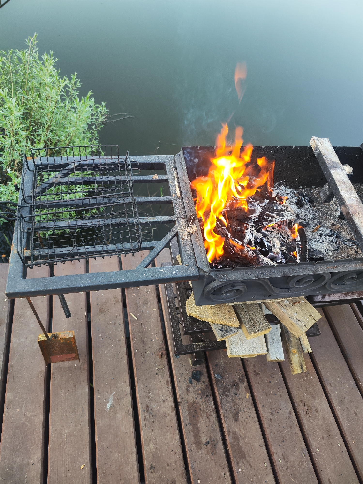 The image shows a black metal grill on a wooden deck. A fire is burning inside the grill, with flames visible. Beside the fire, there are ashes and unburnt wood. A metal grill basket sits on an adjacent platform of the grill. Under the grill, there are several pieces of wood lying on the deck. In the background, there is a body of water and a green bush. The location is Chaikovsky, Russia.