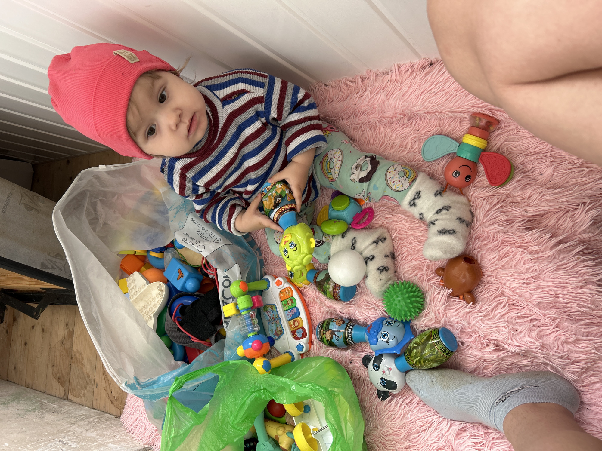 A baby is pictured sitting on a pink fluffy rug, surrounded by a multitude of colorful toys, engaged in playtime. The scene appears to be indoors, possibly in a home in Chaikovsky, Russia, with a light-colored wooden floor and white paneled walls visible in the background.
The primary subject is a young child, likely a toddler, wearing a bright pink beanie, a striped long-sleeved shirt (red, blue, and white stripes), and light-colored pants adorned with donut and rainbow patterns. The child is holding a multi-colored toy, possibly a rattle or shaker, and looking directly towards the camera with a curious, neutral expression.
To the child's left is a large, clear plastic bag overflowing with various toys, including building blocks, bright plastic shapes, and other items. A smaller, bright green plastic bag also holds toys and is partially visible. Spread across the pink rug around the child are numerous other toys: rattles, shakers with animal faces (like a panda, lion, and monkey), a bright green spiky ball, a red-winged caterpillar-like toy with a smiley face, a white ball, and a small toy piano/musical instrument.
An adult's leg and foot, wearing a light blue sock with a visible hole in the heel, are partially visible on the right side of the frame, resting on the same pink rug, suggesting an adult is nearby, possibly supervising or participating in the play.
Visible text includes Cyrillic script on the clear plastic bag, with words such as "ТОЛЬКО ДЛЯ" (ONLY FOR), "БОЛЬШОЕ" (BIG), and fragments like "ЕДИНЫЙ CУПЕРНОЙ" (SINGLE SUPERIOR). The overall atmosphere is one of domestic comfort and playful activity.