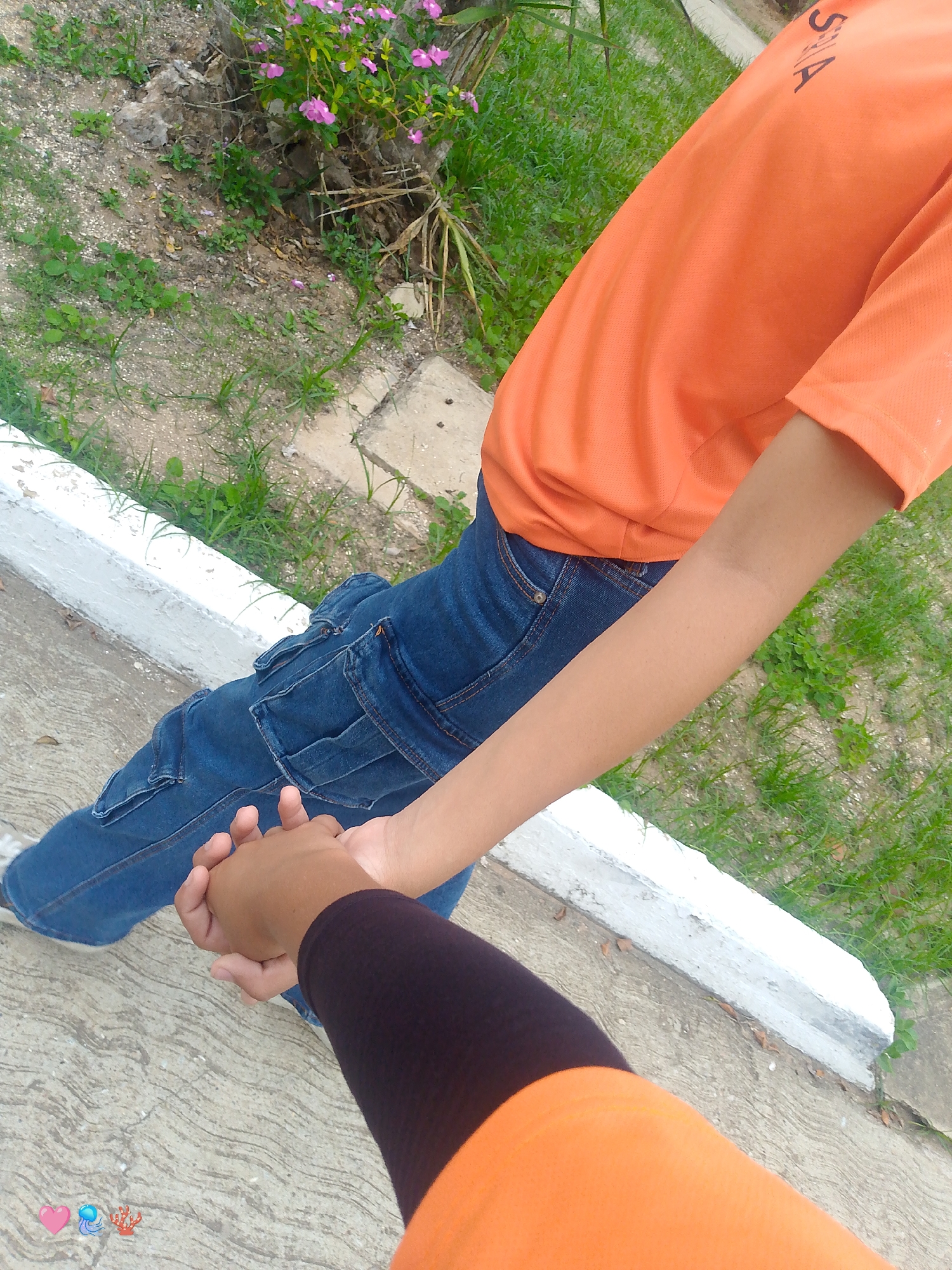 The image shows a close-up, over-the-shoulder view of two people holding hands. One person is wearing an orange t-shirt with the text "SIA" visible on the back. They are also wearing blue denim cargo pants. The other person is wearing a black arm sleeve and an orange top, suggesting they are also wearing an orange shirt underneath. They are holding hands, with the person in the black arm sleeve's hand on top.
The setting appears to be outdoors, with a paved path in the foreground and grass and some pink flowers in the background, hinting at a park or natural area. A white concrete curb runs along the edge of the path. The lighting suggests it is daytime. The image is cropped tightly on the individuals' hands and torsos, focusing on the connection between them. The overall mood conveyed is one of connection and intimacy, with the act of holding hands being the central focus. There are also small emoji icons of a pink heart, a jellyfish, and coral at the bottom left of the image. The location context provided is Playa El Agua, Venezuela.