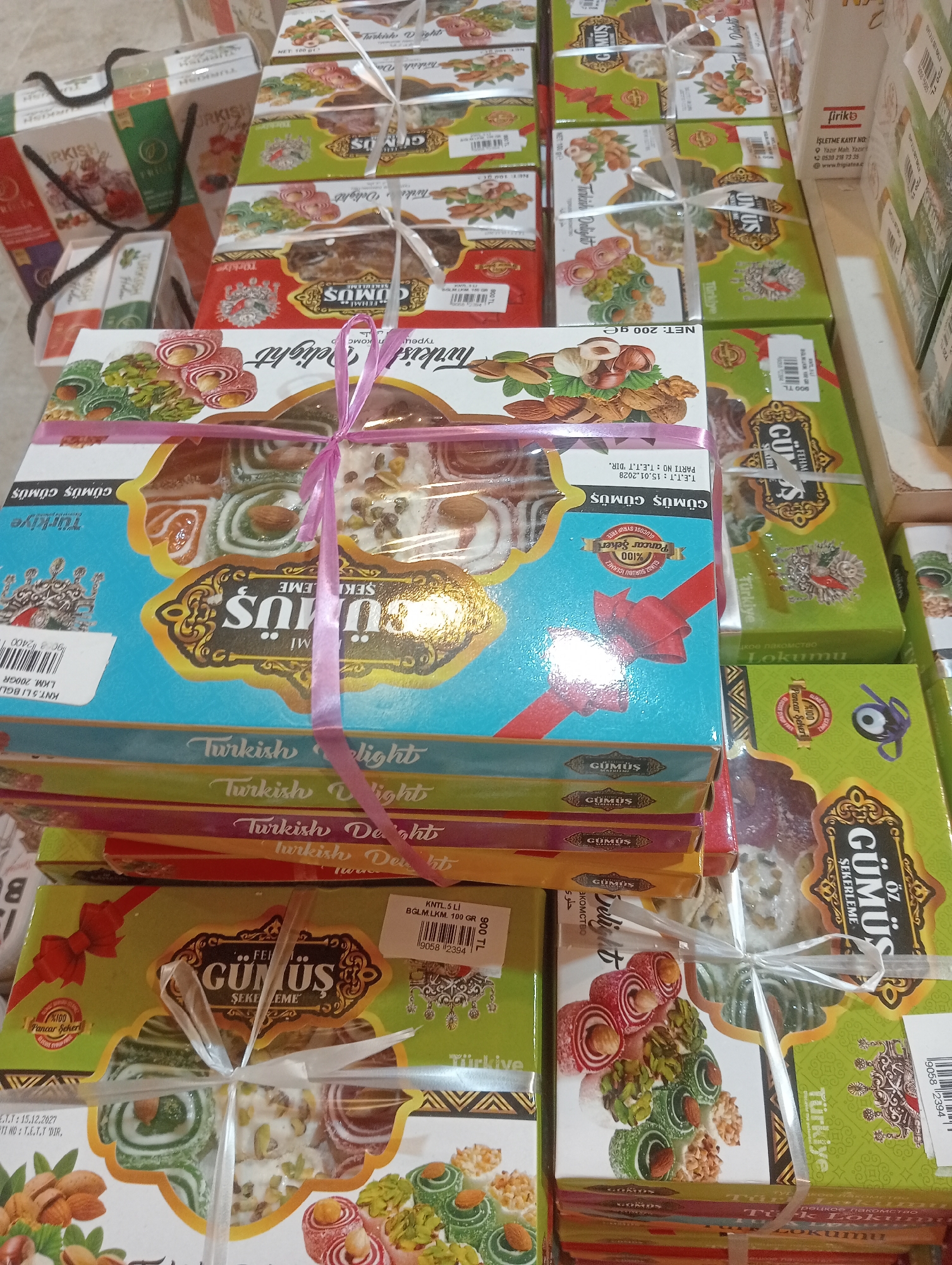The image shows a stack of Turkish delight boxes, predominantly in shades of green and blue, with intricate gold and floral designs. Some boxes are tied with white and pink ribbons. The brand name "GÜMÜŞ" appears multiple times on the packaging, along with the words "Turkish Delight" in English and Turkish. Some boxes also feature images of nuts and the confection itself. The scene appears to be an indoor setting, likely a shop or a market stall, given the stacked nature of the products. The lighting suggests artificial illumination. There are no people visible in the frame. The setting is identified as being in the city of Korkuteli, Turkey, though no specific landmarks or environmental cues within the image directly confirm this location.