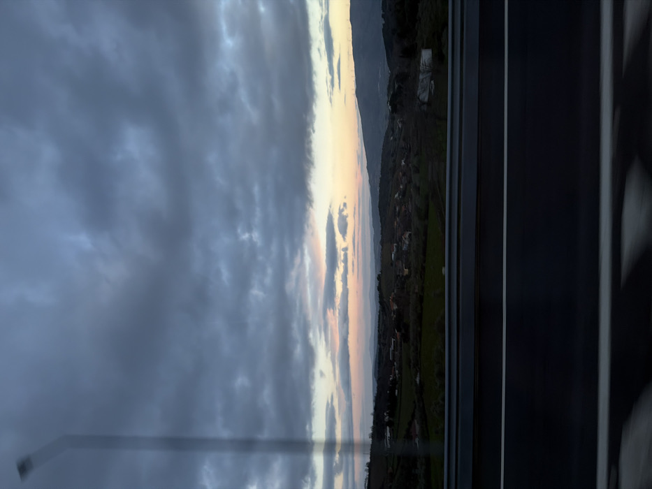 The image displays a landscape scene viewed through a window, captured during either sunrise or sunset. The perspective suggests the photograph was taken from inside a structure, possibly a building or a vehicle, looking outwards.
The upper portion of the image is dominated by a vast and dramatic sky, characterized by heavy, dark grey and blue-grey clouds. These clouds are thick and appear somewhat turbulent. Towards the middle and right side of the sky, the clouds thin or break, revealing a strip of softer light. This area exhibits a beautiful blend of pale yellow, soft orange, and subtle pink hues, indicative of the sun being low on the horizon, either rising or setting. Smaller, more defined cloud formations are also visible within this lighter band of sky.
Below the horizon, the landscape unfolds with gently rolling hills and mountains. The foreground and mid-ground hills are covered in lush green vegetation, possibly agricultural fields or pastures, and are dotted with clusters of small buildings or houses, suggesting a rural village or settlement. Further in the distance, mountain ranges appear darker and more subdued in color, partially obscured by the ambient light conditions and distance.
On the far right side of the image, a prominent dark, vertical band extends from top to bottom, which is clearly a window frame. This frame, along with a narrower vertical line to its left, creates a visual barrier, framing the landscape view. There are also hints of reflections or internal structures within the dark frame. A faint, translucent, elongated, light-colored rectangular shape, possibly a reflection or a part of the window's structure, extends diagonally from the lower left corner towards the center-left sky. Minor smudges or streaks are also discernible on the window's surface, particularly in the lower-left area.
The overall mood of the image is serene yet somewhat dramatic, conveyed by the contrasting light and dark elements of the sky and landscape. No text is visible in the image.