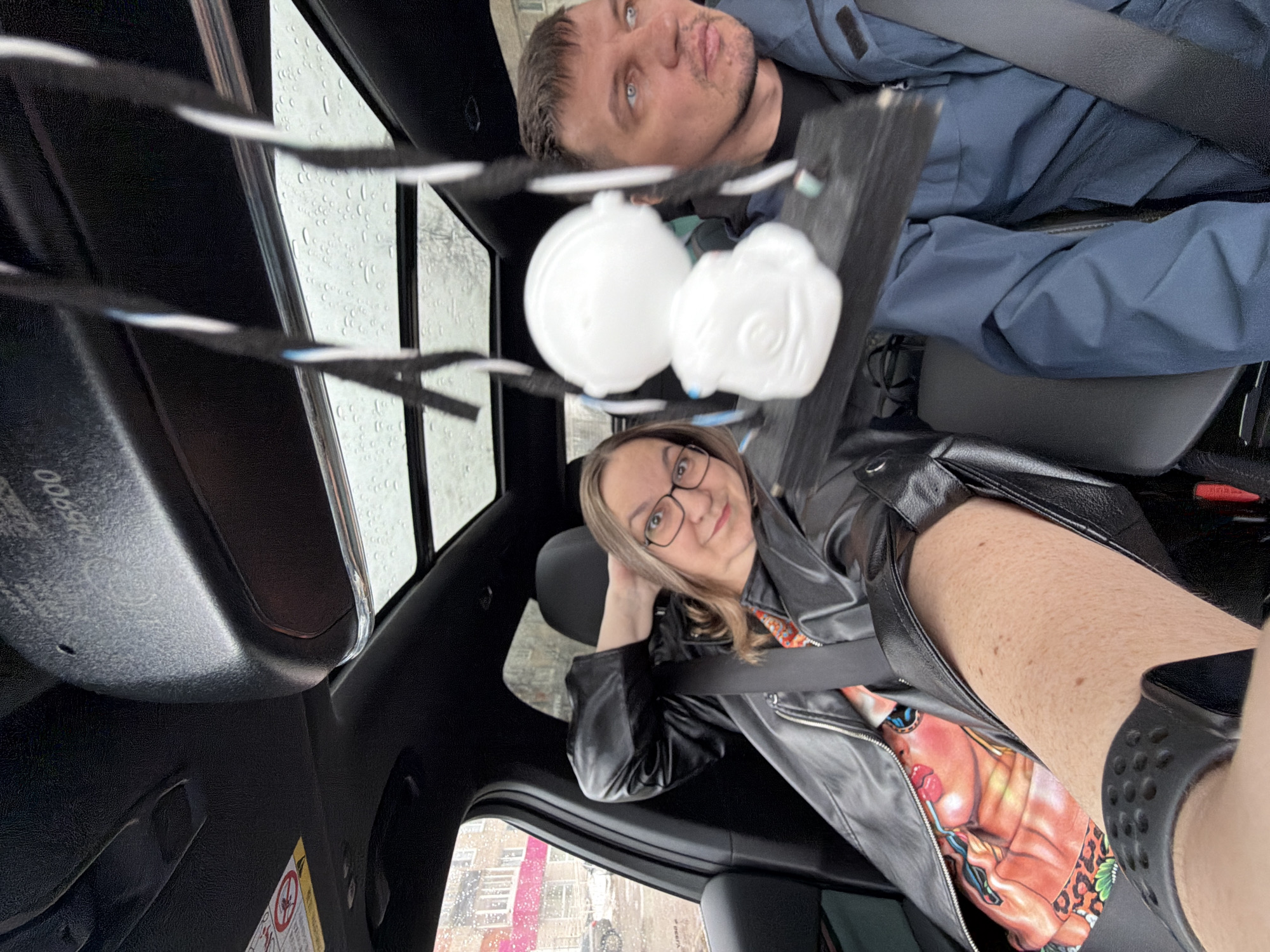 A selfie photo taken inside a car. Two people are visible. A woman wearing a black leather jacket, a patterned top, glasses, and a black smartwatch on her left wrist is seated, facing the camera. A man wearing a blue jacket is above her, also facing the camera. Two white, decorative objects are suspended near the car's ceiling. Rain is visible on the car windows. Through the windows, a building with pink and red is visible. Based on location information embedded in the metadata, the photo was taken in Chaikovsky, Russia.