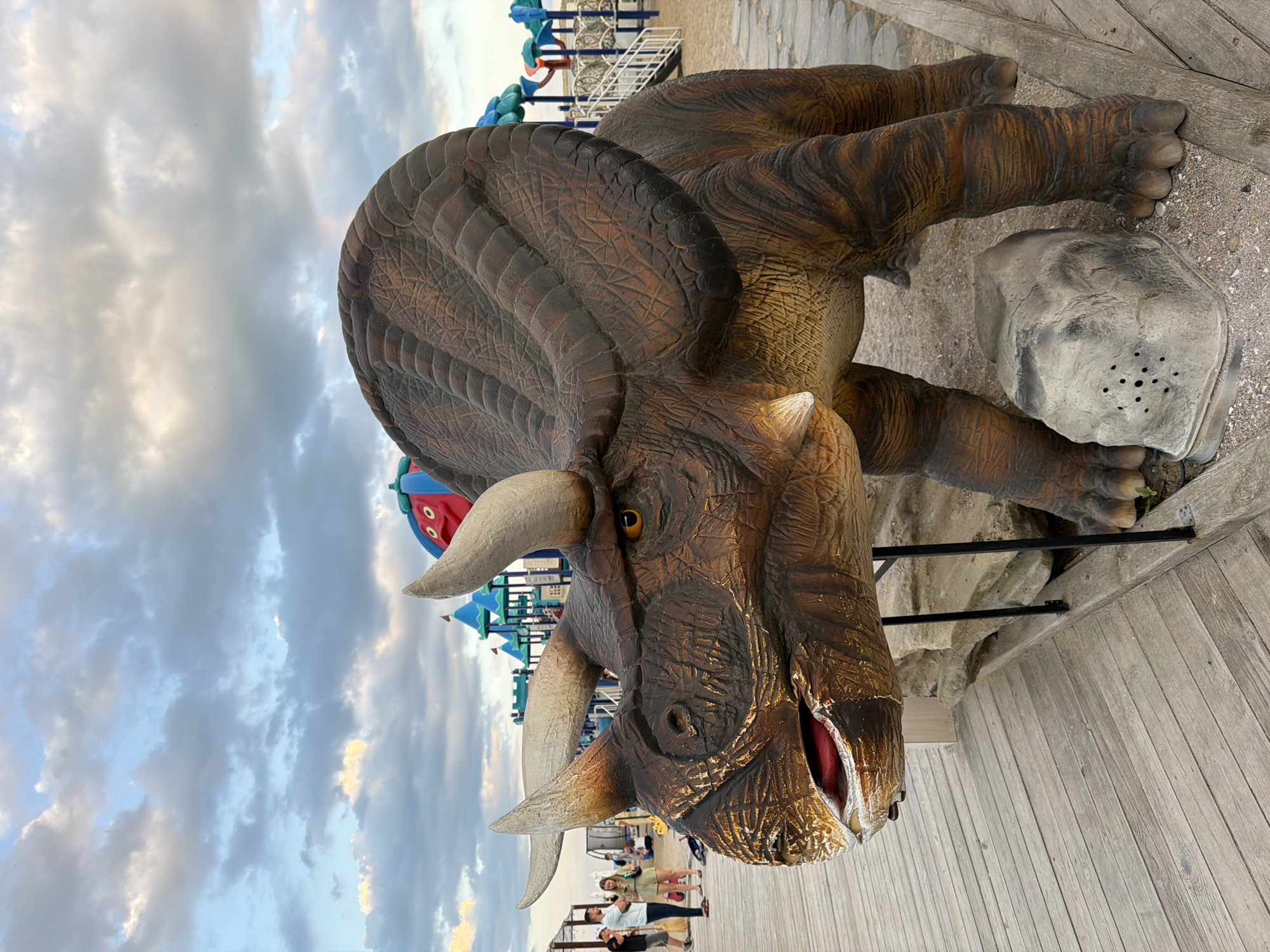 The image depicts a life-sized dinosaur sculpture, specifically a Triceratops, positioned on a wooden boardwalk. The sculpture is detailed, with brown textured skin and prominent horns. In the background, there is a playground with colorful structures, including slides and climbing frames, suggesting an outdoor recreational area. Several people are visible in the distance, some walking and others sitting, appearing to be on a beach or sandy area. The sky is cloudy with patches of blue, indicating it is likely late afternoon or early evening. The location is identified as Fontanka, Ukraine.