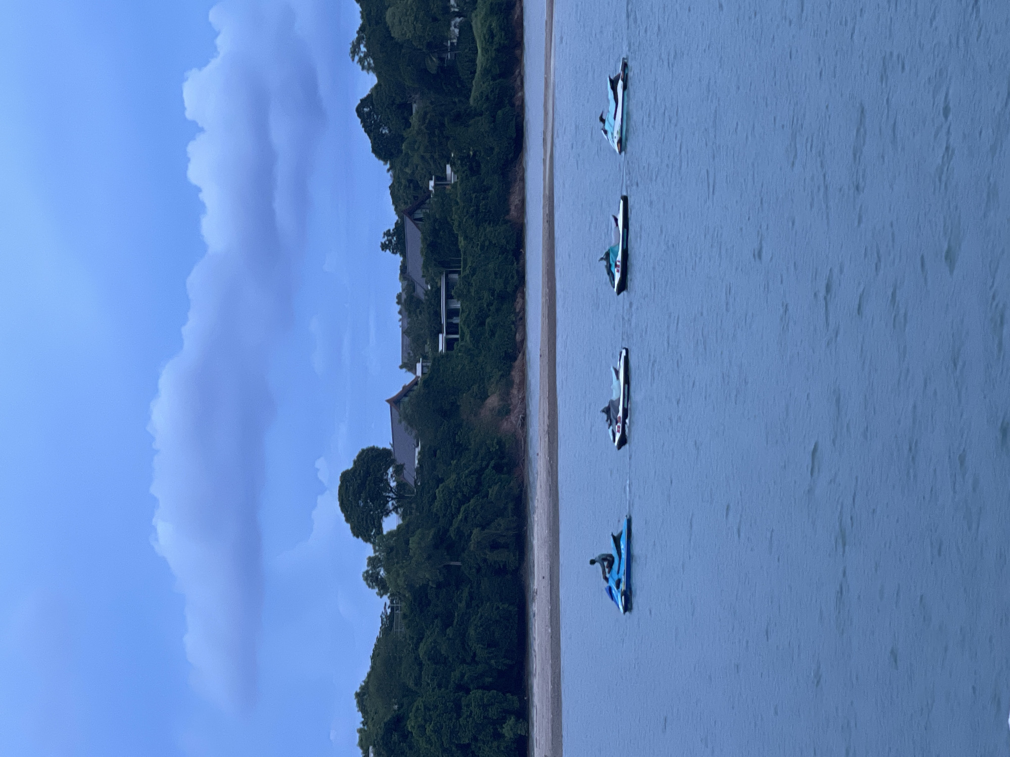 A scenic view of the sea in Pa Khlok, Thailand, showcases a line of four jet skis gliding across the calm, blue water. The lead jet ski is a vibrant blue and has a person riding it. Behind it, three other jet skis, two primarily white with turquoise accents, and one with black and white accents, follow in a staggered formation. The backdrop features a lush, green hillside densely covered with trees, with several modern houses and villas nestled amongst them. A thin strip of sandy beach separates the water from the hillside. The sky above is a deep blue, with soft, elongated clouds adding a dramatic touch to the twilight atmosphere. The image captures a tranquil moment of recreational activity against a beautiful natural and residential landscape.