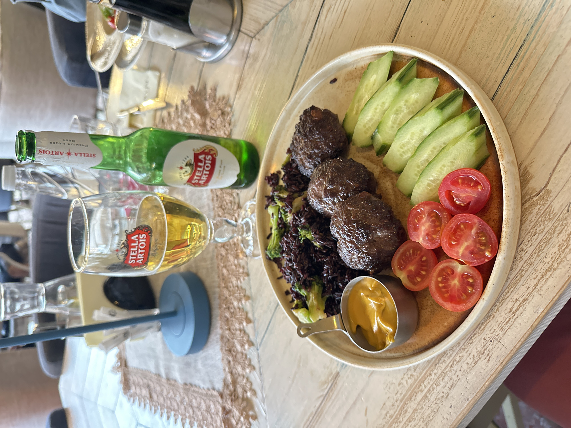 The image shows a partially eaten plate of food and a bottle and glass of Stella Artois beer on a table. The plate contains three meatballs, a side of dark rice or grain with greens, slices of cucumber, sliced cherry tomatoes, and a small dish of mustard. The bottle of beer is green and appears to be partially full. The glass of beer is about half-full. The table has a light wood grain finish and a textured linen runner. Although the precise location cannot be definitively determined from this single image, the subject matter is consistent with restaurant dining experiences in urban locations.