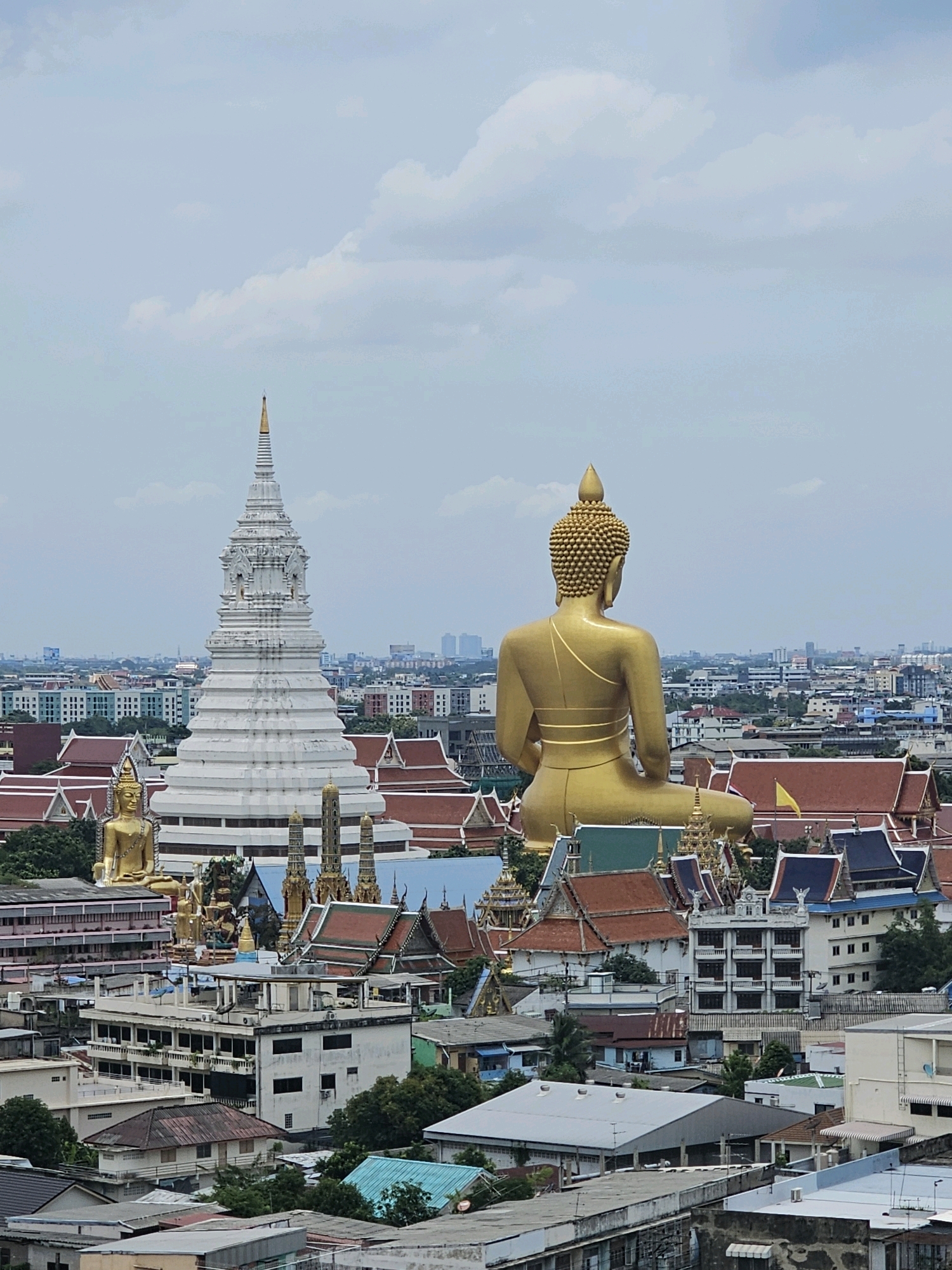 This is a medium-shot, slightly high-angle, full-color, vertical photograph taken on an overcast day in Bangkok, Thailand, featuring a panoramic view of a city skyline dominated by a massive golden Buddha statue and a large white stupa. The golden Buddha, seated in a meditative pose, faces away from the viewer, its back and shoulders filling the right side of the frame. It is positioned behind and above a complex of Thai temples with distinctive multi-tiered, ornate roofs.
To the left of the golden Buddha, a prominent white stupa with intricate carvings and a golden spire rises majestically. It stands out against the hazy, pale blue sky dotted with soft white clouds. Smaller golden Buddha statues and temple spires are visible in the midground, interspersed with traditional Thai temple architecture.
The foreground and midground are filled with a dense urban landscape of low-rise buildings with a variety of roof styles and colors, including terracotta tiles and corrugated metal. Many of these buildings appear to be residential or commercial structures. In the distance, the urban sprawl continues, with taller buildings and a more modern cityscape visible against the horizon. The overall impression is one of a vibrant city where ancient religious structures coexist with modern urban development.