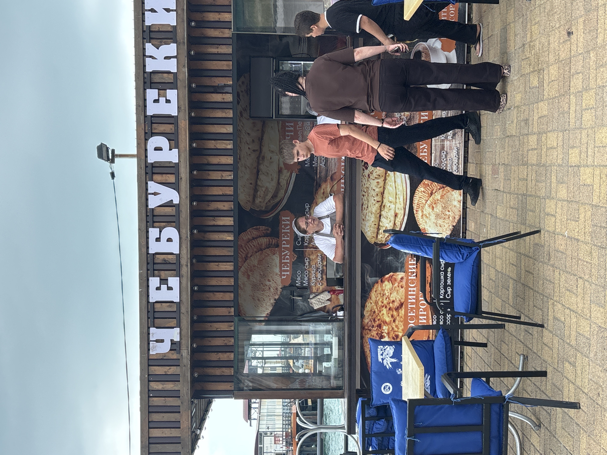 The image depicts an outdoor food stall in Sochi, Russia, specifically a cheburek vendor. The stall has a wooden facade with the word "ЧЕБУРЕКИ" (Chebureki) prominently displayed in large white letters across the top. Below the sign, a window reveals a woman in a white uniform serving customers. The counter displays images of various chebureki and other baked goods. In front of the stall, several people are visible. A young man in an orange t-shirt and black pants is walking away from the counter, while a woman with braided hair and dark clothing stands beside him, facing away from the camera. A younger boy in a black t-shirt and dark pants is on the other side of the woman, also facing away. To the left of the stall, there are blue chairs and a table, suggesting an outdoor seating area. The ground is paved with yellow bricks, and in the background, a body of water and some buildings can be seen. The sky is overcast, indicating potentially cloudy weather.