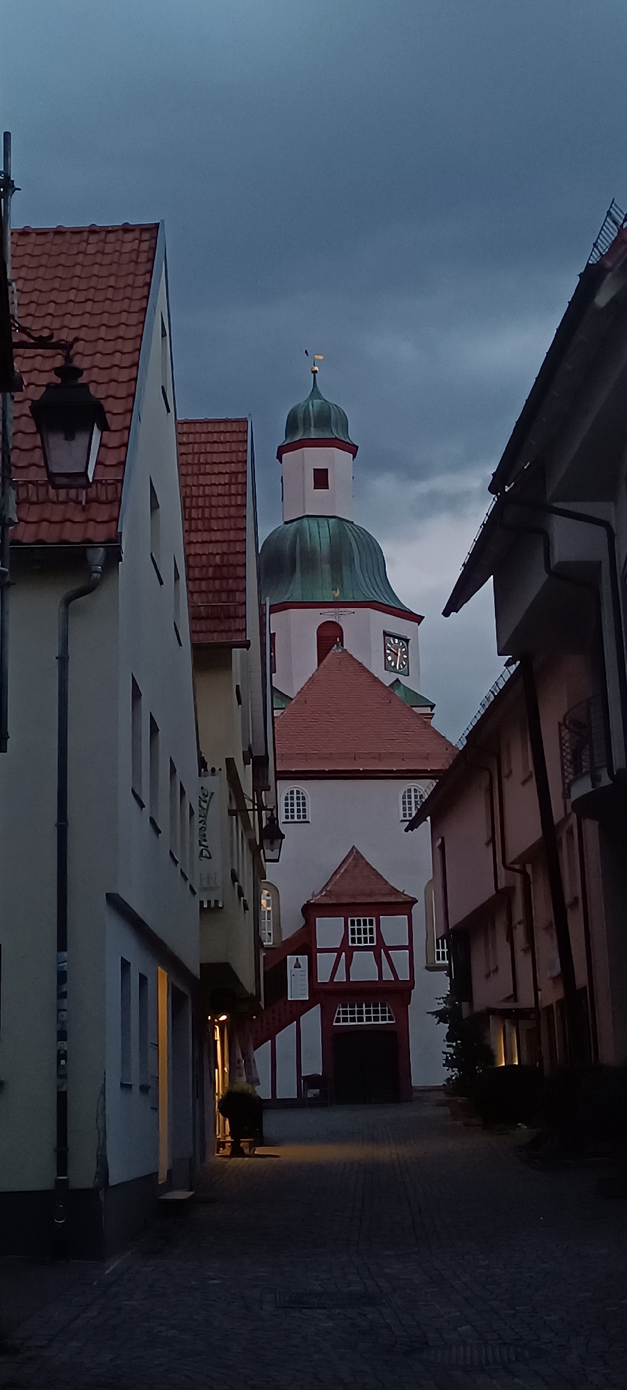 The image captures a street scene in Winnenden, Germany, featuring a prominent church tower with a green dome and a clock. The perspective is from a narrow cobblestone street lined with buildings on both sides. On the left, a white building with a red-tiled roof and a black lantern is visible, with an awning extending over an open doorway. A sign with the word "Dressure" is mounted on this building. On the right, other buildings with pitched red roofs and dark facades contribute to the confined feel of the street. The church tower, with its white facade, red-trimmed windows, and green dome topped with a cross, is the central focal point. A red-tiled roof section with white-framed windows is situated in front of the church entrance, and an external staircase leads up to a section of the church with a half-timbered structure. The sky is overcast and dark, suggesting twilight or evening. A single figure is dimly visible in the light emanating from a doorway on the left side of the street, appearing to be pushing a trolley.