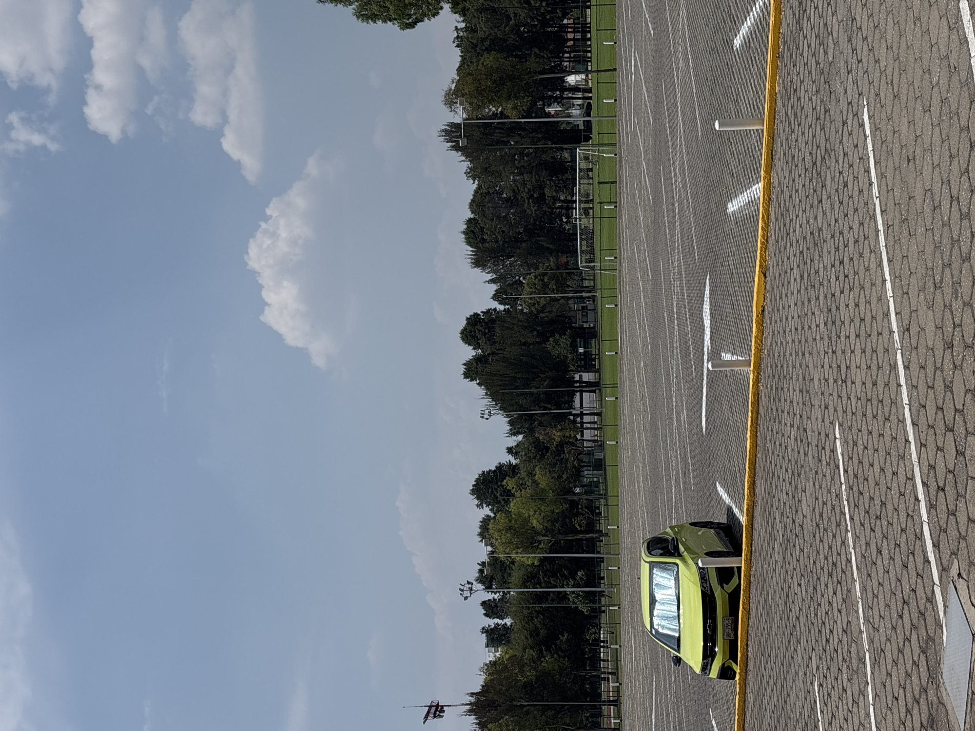 The image depicts a lime green Chevrolet Bolt parked in a paved parking lot with white lines and a yellow curb. The parking lot is adjacent to a green space with trees and what appears to be a soccer field with goals. The sky is blue with scattered white clouds. A flag pole is visible in the distance. The license plate on the car reads "BBP-755-B", indicative of license plates issued in the State of Puebla, Mexico. Given the presence of the soccer field and the parking lot layout, this location could be in or near a sports complex in San Andrés Cholula, Mexico.