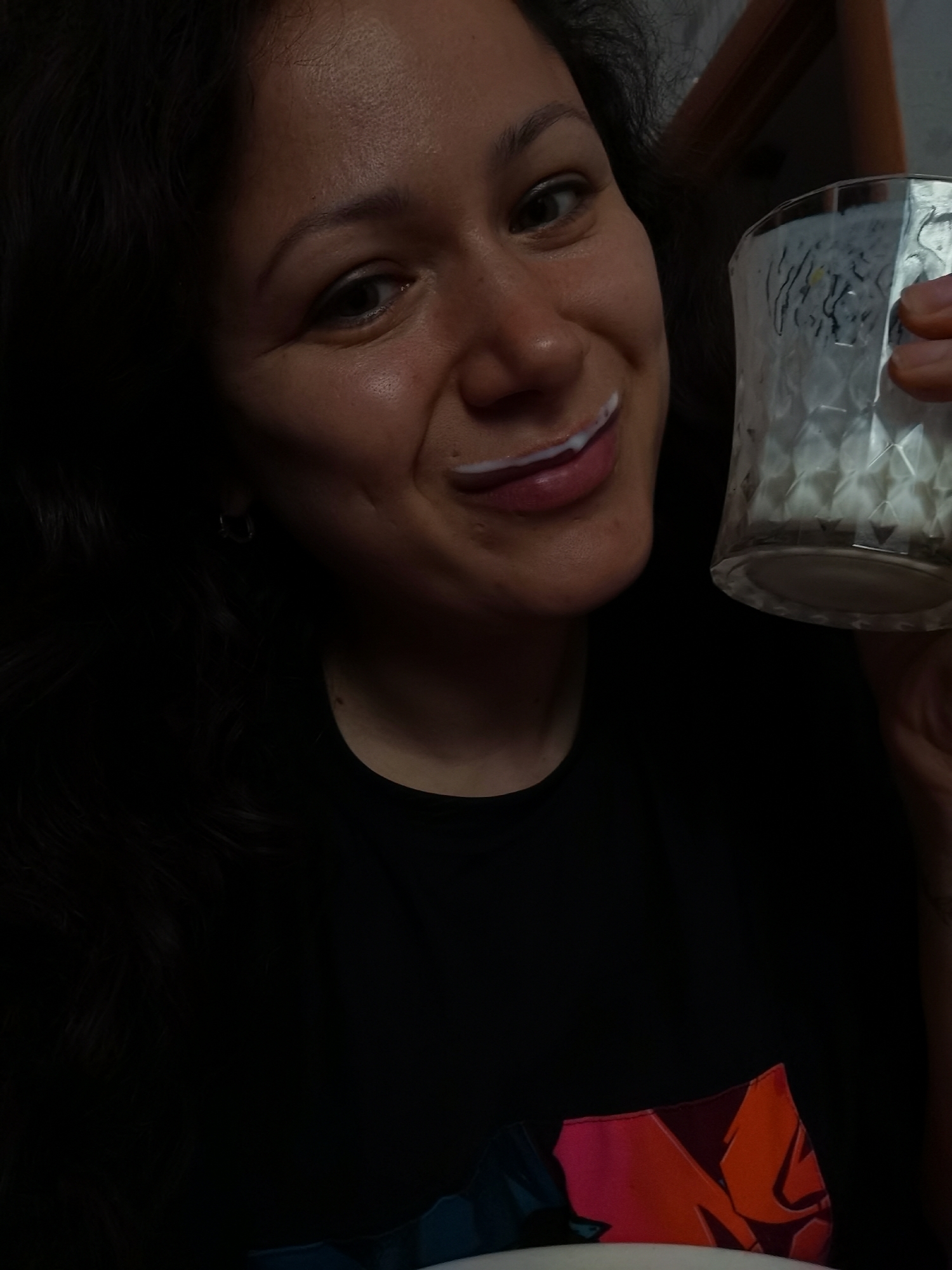 A close-up shot captures a woman with dark, wavy hair and a faint milk mustache. She is looking towards the camera, holding a glass of milk in her right hand. Her lips are slightly parted, revealing white teeth. The background is dark, with hints of brown and orange furniture visible, suggesting an indoor setting. The woman is wearing a black t-shirt with a graphic design on it. The image appears to be taken in Chaikovsky, Russia, based on the provided context.