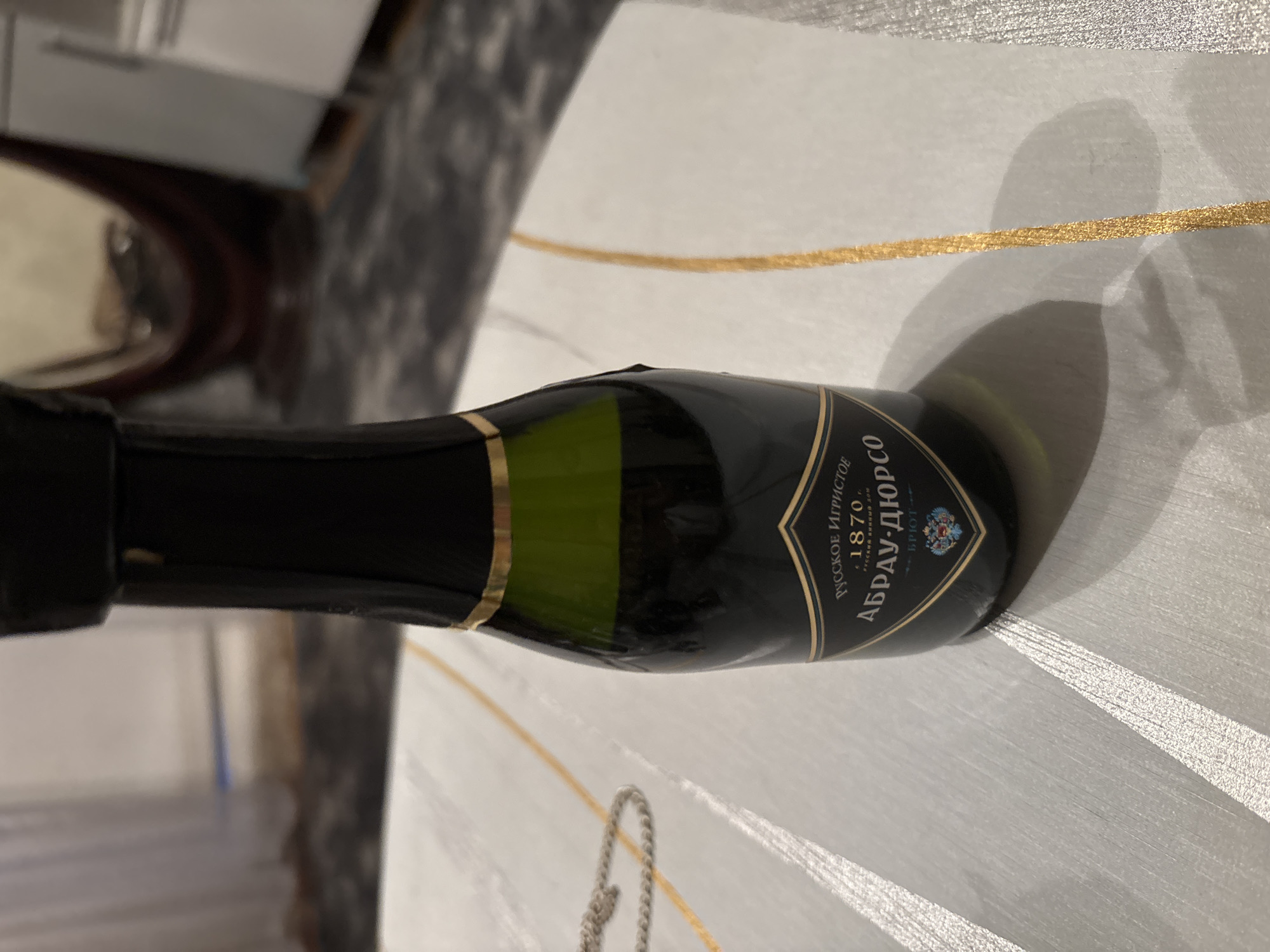 A miniature bottle of sparkling wine, labeled in Russian as "АБРАУ-ДЮРСО," sits on a textured, light-colored surface. The bottle is dark green glass, topped with a black capsule and a gold band. The label is black with gold lettering and features a depiction of a coat of arms. The surface it rests on has gold and silver linear decorations. The background is blurred, showing hints of an indoor environment with curtains and furniture. The scene is likely indoors in Chernogorsk, Russia.
