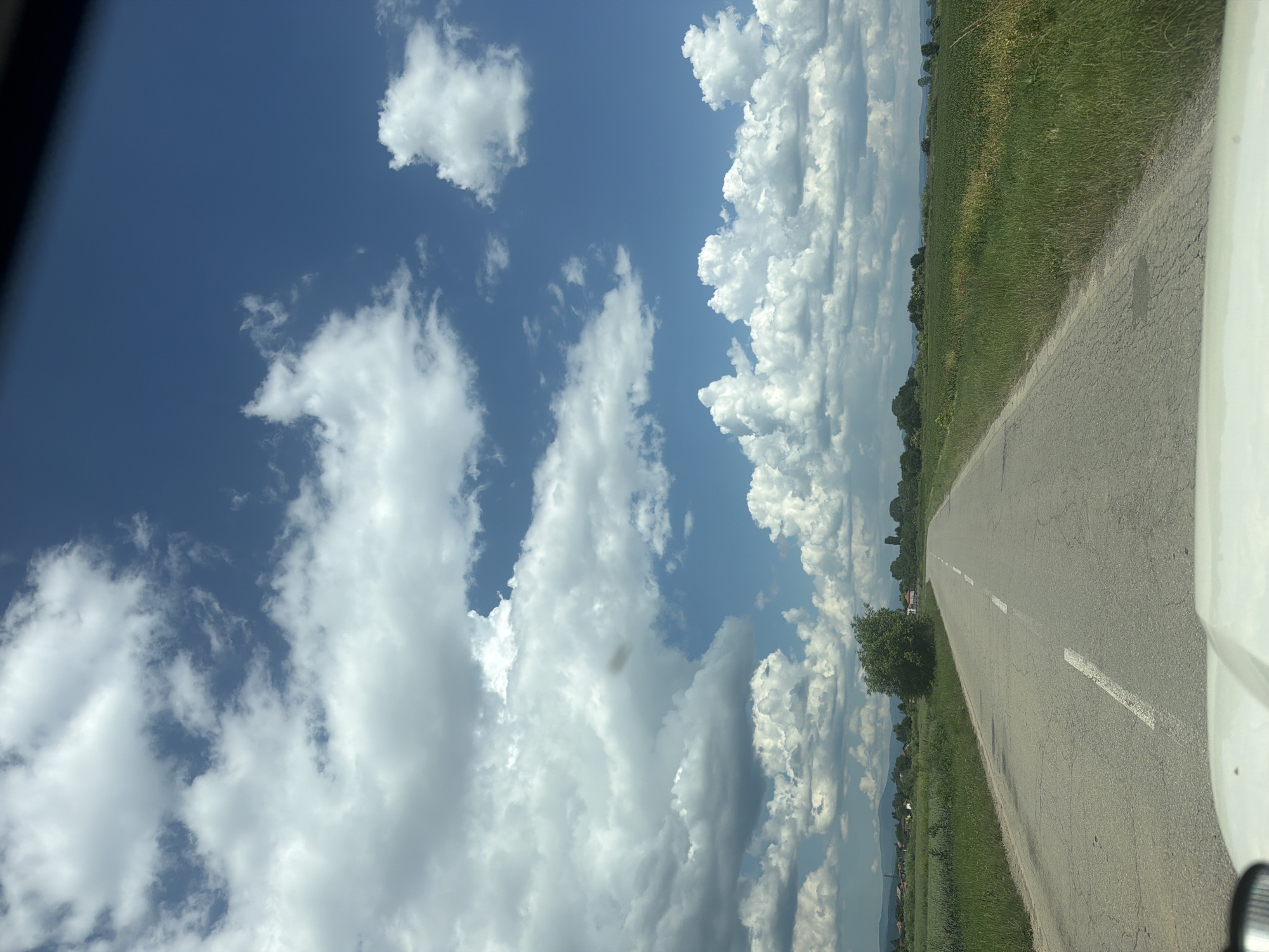 The image captures a paved road in Musachevo, Bulgaria, leading through a rural landscape under a dynamic sky filled with cumulus clouds. The road, marked with a white dashed line, is bordered by lush green fields on both sides. In the distance, trees and a few houses are visible, with rolling hills forming the horizon. The overall scene depicts a serene, daytime view of the Bulgarian countryside.