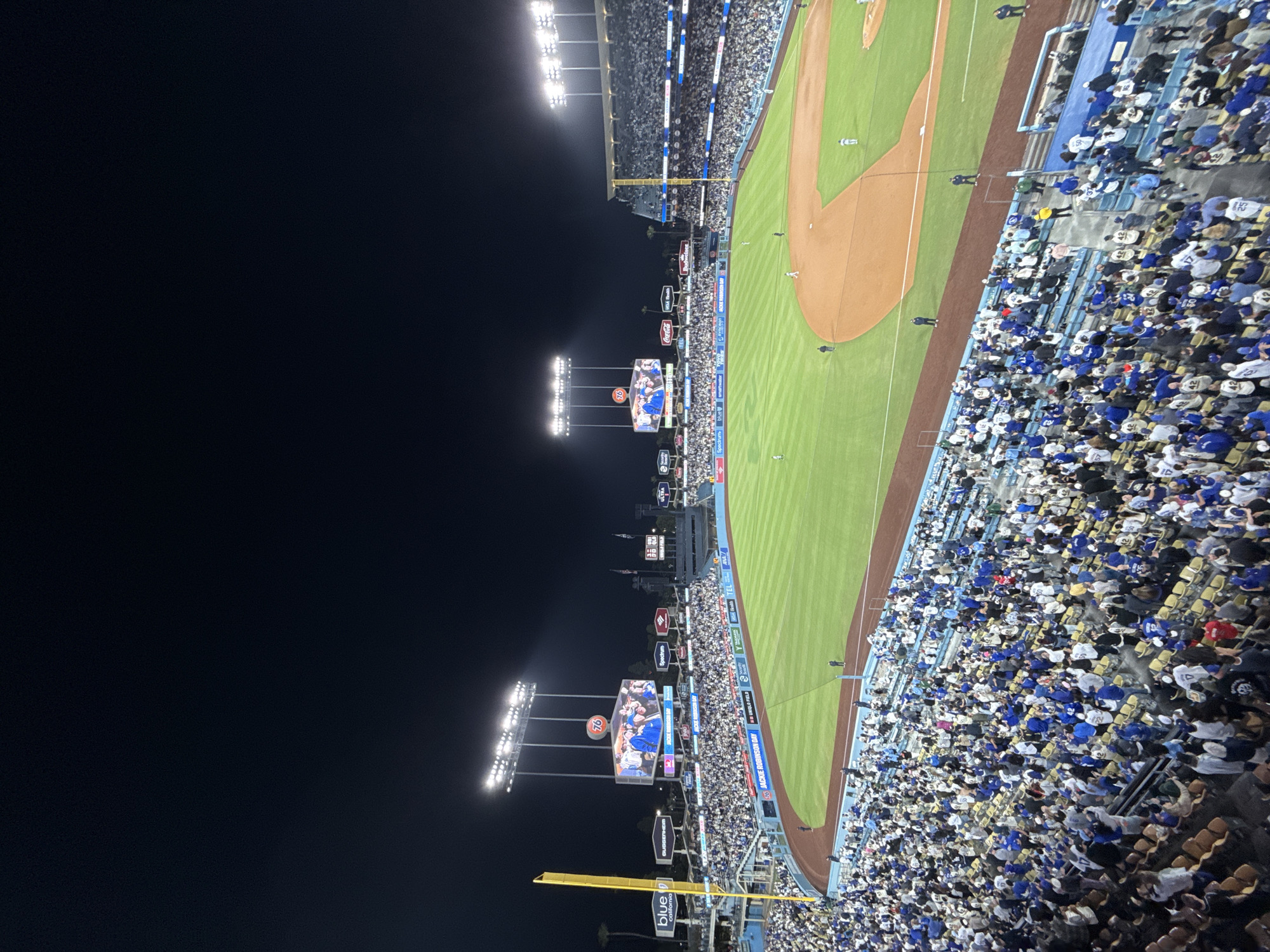 The image depicts a nighttime baseball game at Dodger Stadium in Los Angeles, USA. The stadium is filled with a large crowd of spectators, many of whom are wearing blue attire. The field is illuminated by stadium lights. The center field video board is showing a Jackie Robinson tribute on the anniversary of his major league debut. Various sponsor logos are visible around the stadium, including those for Coca-Cola, Spectrum, and Blue California.