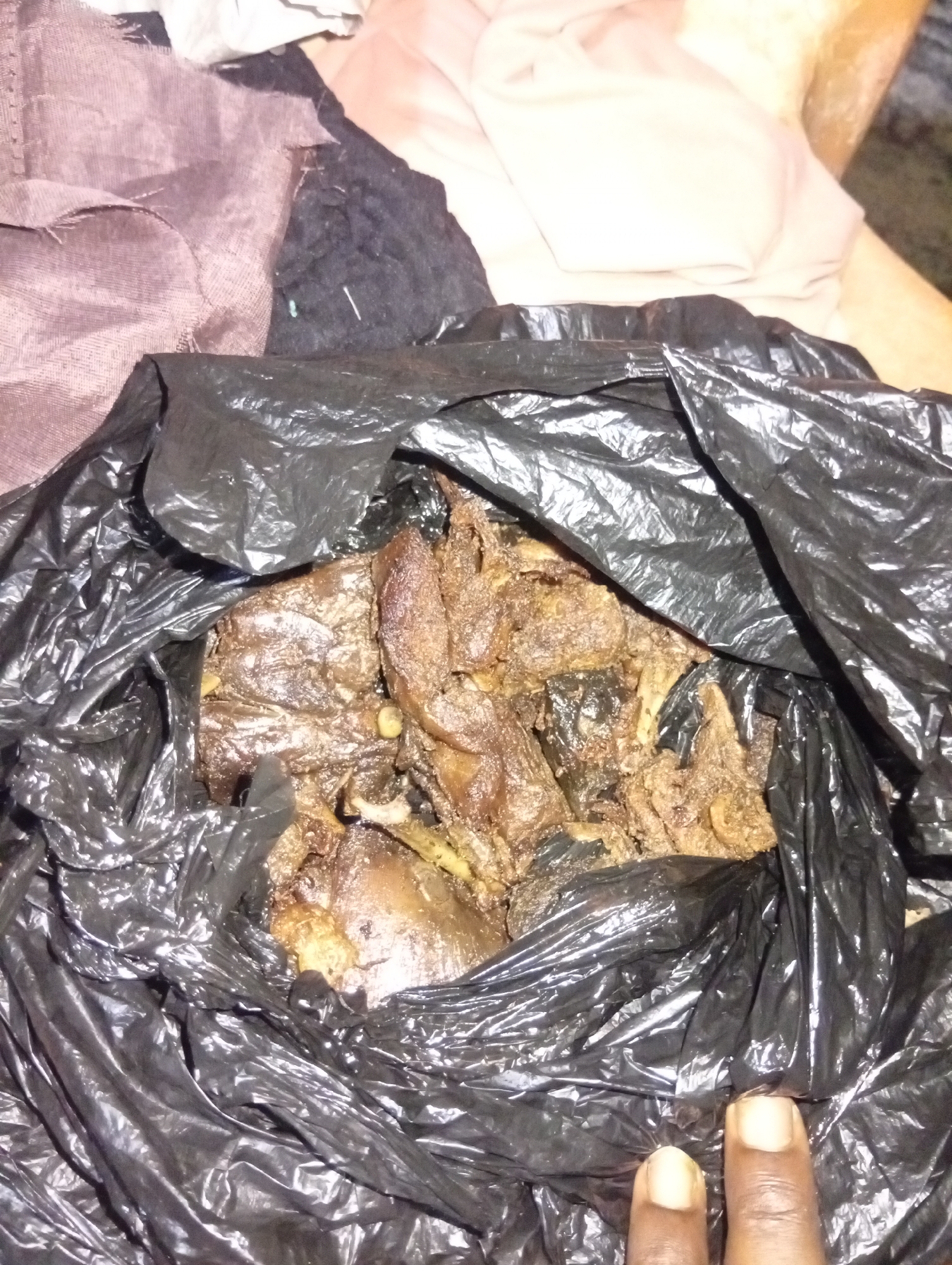 A black plastic bag containing cooked meat and bones sits on a surface. Two fingers are visible in the bottom right of the frame. The background includes fabric in various colors. The location is Aviation Quarters, Nigeria.
