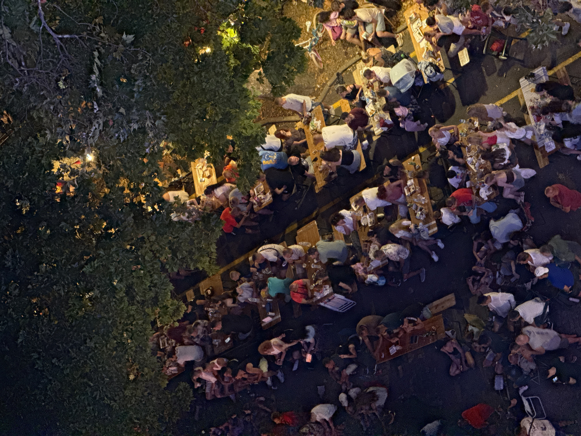 This aerial photograph captures an outdoor dining scene at night in Burgas, Bulgaria, possibly at a restaurant or beer garden. Long wooden tables are arranged in rows, occupied by numerous people seated and standing. The atmosphere appears lively, with groups of individuals engaged in conversation and enjoying their meals or drinks. The lighting is dim, with some artificial lights and the glow of string lights or lamps illuminating the area and creating dappled light through the trees. The environment is characterized by trees providing shade and a dark asphalt or paved ground. Various objects are visible on the tables, including plates, glasses, and bottles. A bicycle is parked near one of the seating areas, suggesting a mode of transportation for some attendees. The overall impression is one of a social gathering taking place in a relaxed, open-air setting.