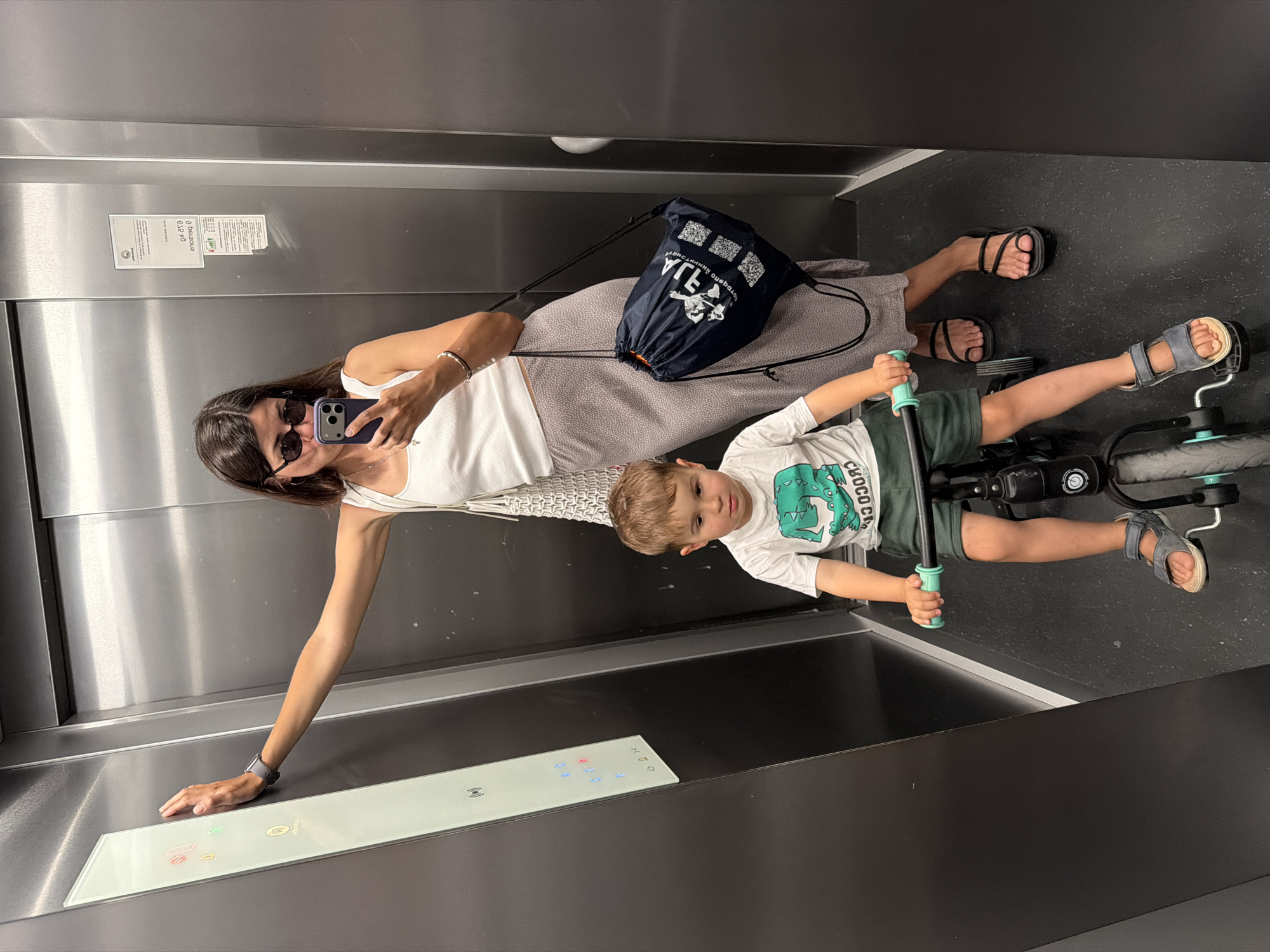 A woman and a young child are in an elevator, presumably in Budva, Montenegro. The woman is taking a selfie with her smartphone. She is wearing sunglasses, a white tank top, and a patterned skirt. She has a dark-colored drawstring bag slung over her shoulder. The child is sitting on a tricycle, wearing a white t-shirt with a dinosaur design and green shorts. The elevator has a control panel with illuminated buttons and stainless steel walls. The floor appears to be dark colored.