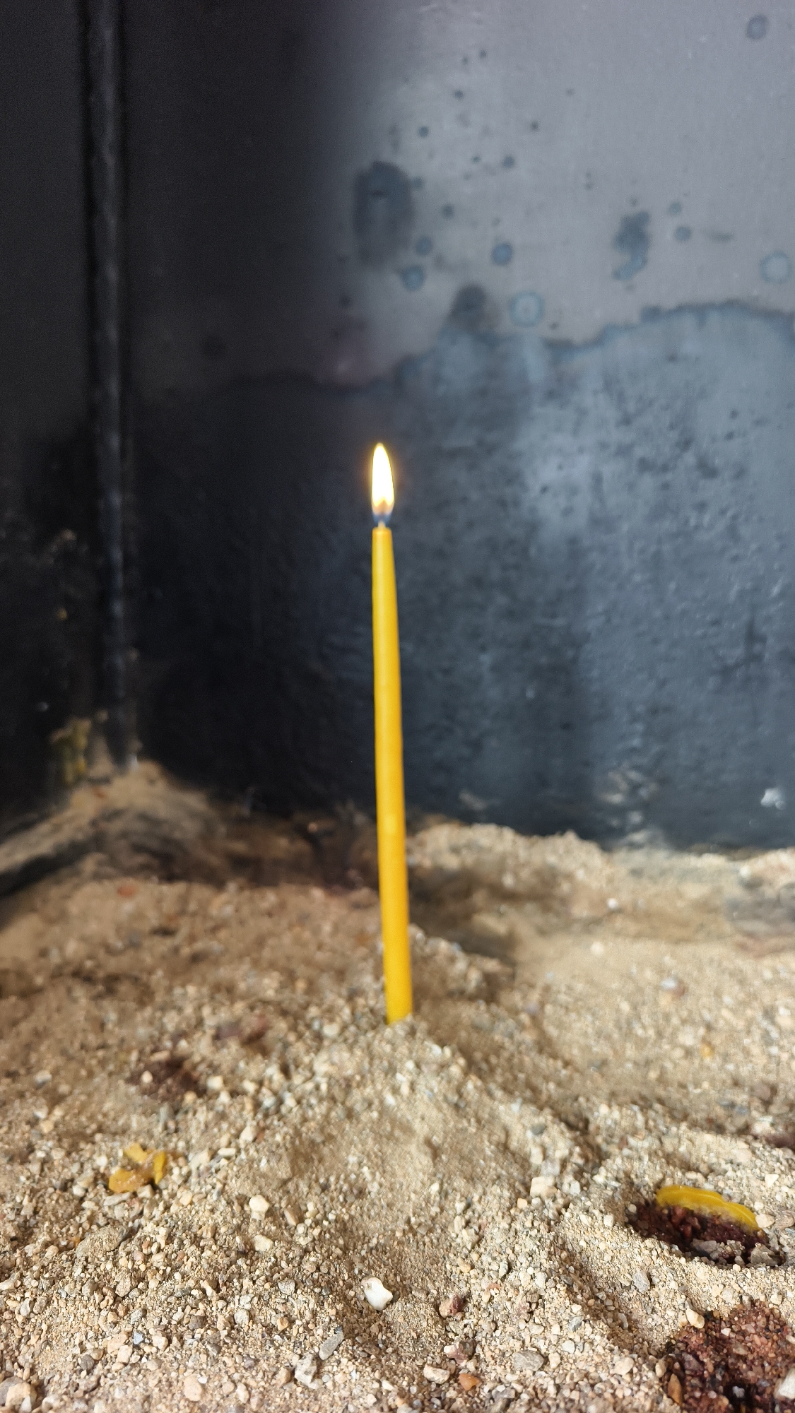 A single yellow candle burns brightly in a sand-filled holder. The candle is positioned vertically in the center of the frame, its flame a vibrant point of light. The sand appears to be composed of fine grains mixed with small pebbles. In the foreground, scattered remnants of what appear to be burnt offerings or debris are visible. The background consists of a dark, rough-textured surface, possibly a wall or the interior of a structure. The overall impression is one of a solemn or religious setting, potentially within the city of Selçuk, Turkey, known for its historical and religious sites. There are no people or animals visible in the image.