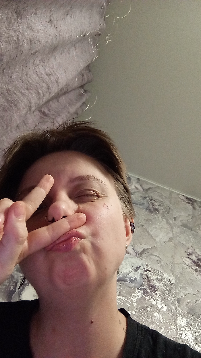 The image captures an upward-angled close-up of an individual indoors, with the location identified as Saratov, Russia. The subject, a person with short, dark brown hair, is positioned centrally, looking towards the upper right. Both eyes are closed, and lips are puckered. The left hand is raised, forming a "peace sign" with the index and middle fingers, held across the lower face, partially obscuring the nose. A metallic ring is visible through the nasal septum. A small, outline-style heart tattoo is present on the right cheekbone below the eye. The right ear exhibits multiple piercings, including a dark-colored, coiled ear cuff or helix piercing. The individual is wearing a dark-colored garment. The upper-left background features a light grey, textured fabric, possibly a curtain, displaying fine reflective fibers. The background to the right and lower frame consists of a wall covered in light-toned wallpaper featuring an abstract, mottled pattern of grey and silverish-white hues. The scene is illuminated by an apparent internal artificial light source.