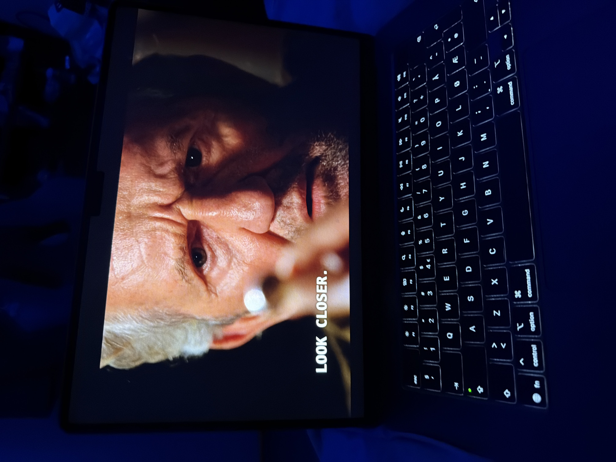 The image shows a close-up of a laptop screen displaying a man's face. The man appears to be middle-aged or older, with graying hair and wrinkles on his face. He has a serious expression and is looking directly at the camera. Text overlaid on the screen reads "LOOK CLOSER." The laptop is open, and the keyboard is visible below the screen, illuminated by backlighting. The scene is dimly lit, with the primary light source appearing to be the laptop screen itself, casting a blueish glow on the surrounding area. The background is dark and out of focus, suggesting an indoor environment. There is no identifiable information in the image to suggest a specific location in Oslo, Norway.