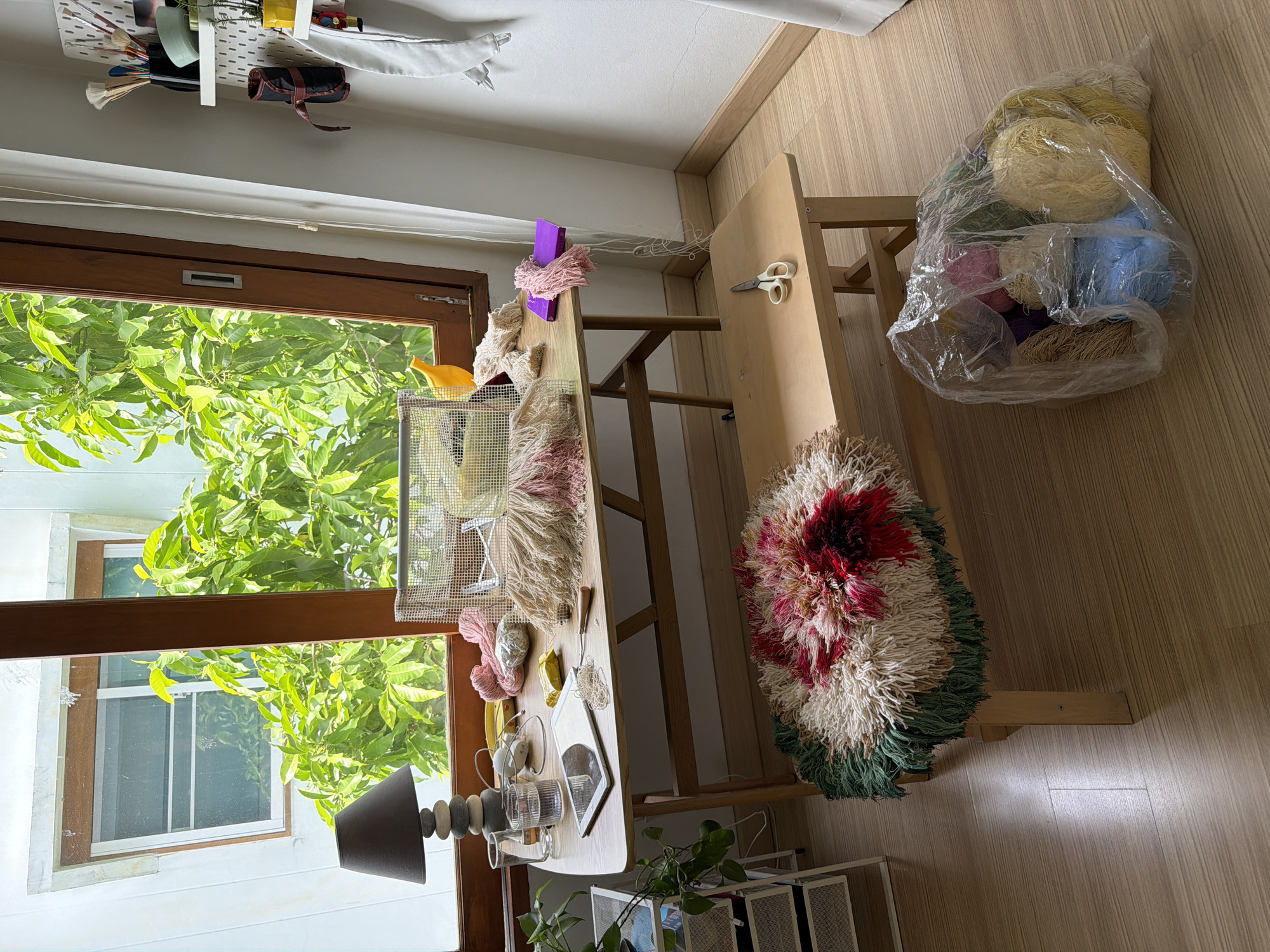 The image shows a crafting space with various materials and tools. A wooden table is set up with yarn, scissors, and a tablet. In the background, a large bag filled with colorful yarn balls is visible. A finished tapestry or rug with a floral design is draped over a chair. The scene appears to be indoors, near a window that looks out onto lush green foliage. The time of day is likely daytime, with natural light filtering through the window. The overall impression is one of a creative workspace, possibly in a home or studio environment. The style of the rug and the abundance of yarn suggest a focus on textile arts or crafts. The location context of Chang Phueak, Thailand, might suggest a warm climate, which would align with the greenery seen outside. There is no visible text in the image.