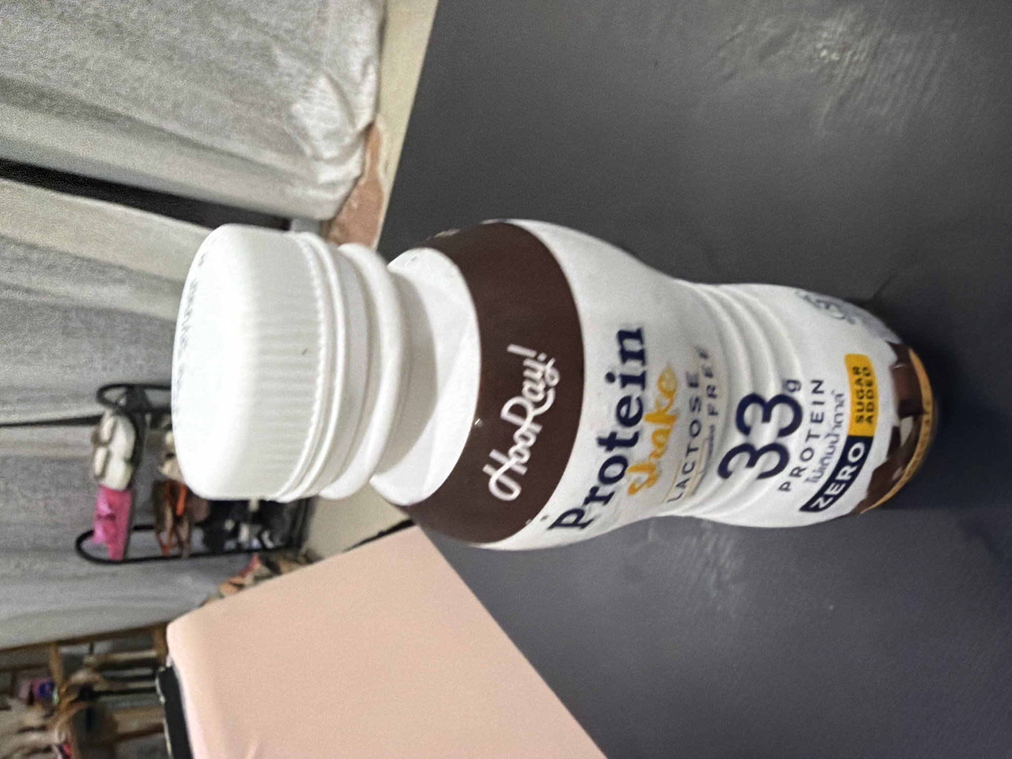 In Baan Mae Nam, Thailand, a brown and white bottle of "HooRay! Protein Shake" sits on a dark, reflective surface, likely a table. The bottle has a white cap and features branding with the text "HooRay!", "Protein Shake", "Lactose Free", "33g PROTEIN", and "ZERO SUGAR ADDED". The background is blurred, showing a pink chair on the left and a white curtain on the right. A shelf unit with shoes and other items is visible in the far background. The lighting suggests an indoor environment.