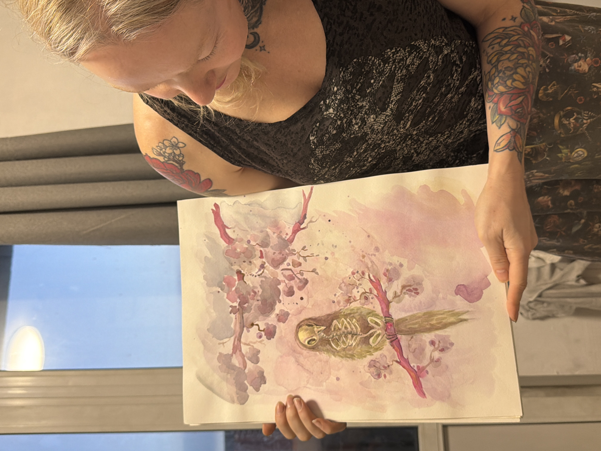A person with tattoos on their arms and neck is holding a watercolor painting. The painting depicts a bird skeleton among cherry blossoms on a light pink and white background. The person is wearing a dark top with a lace-like pattern. Behind the person, there is a window with gray blinds and a view of an indeterminate outdoor scene. There is no indication of the city being Saint Petersburg, Russia.